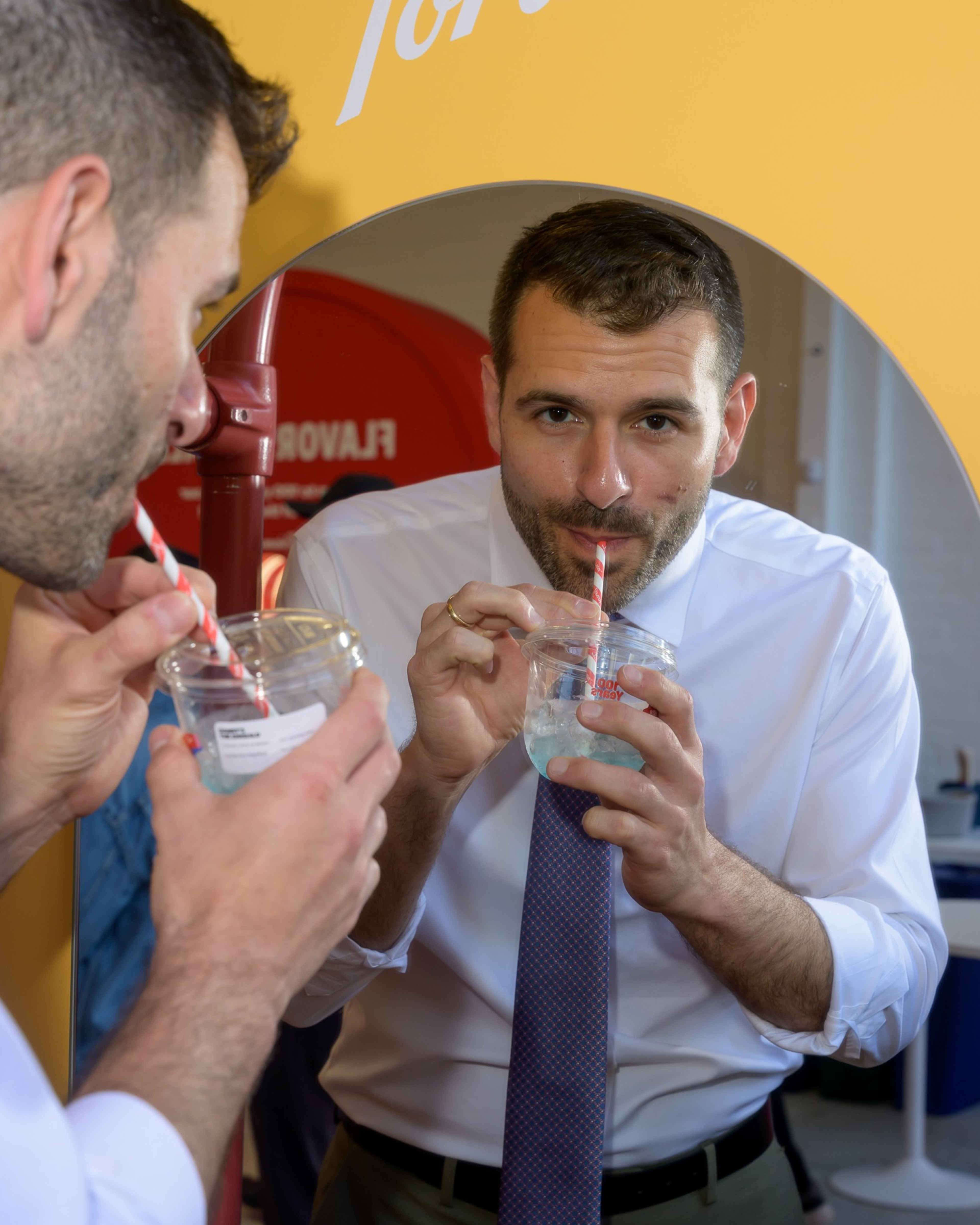 A man in a white shirt and tie drinks a beverage through a red and white striped straw, looking into a mirror reflecting his face.