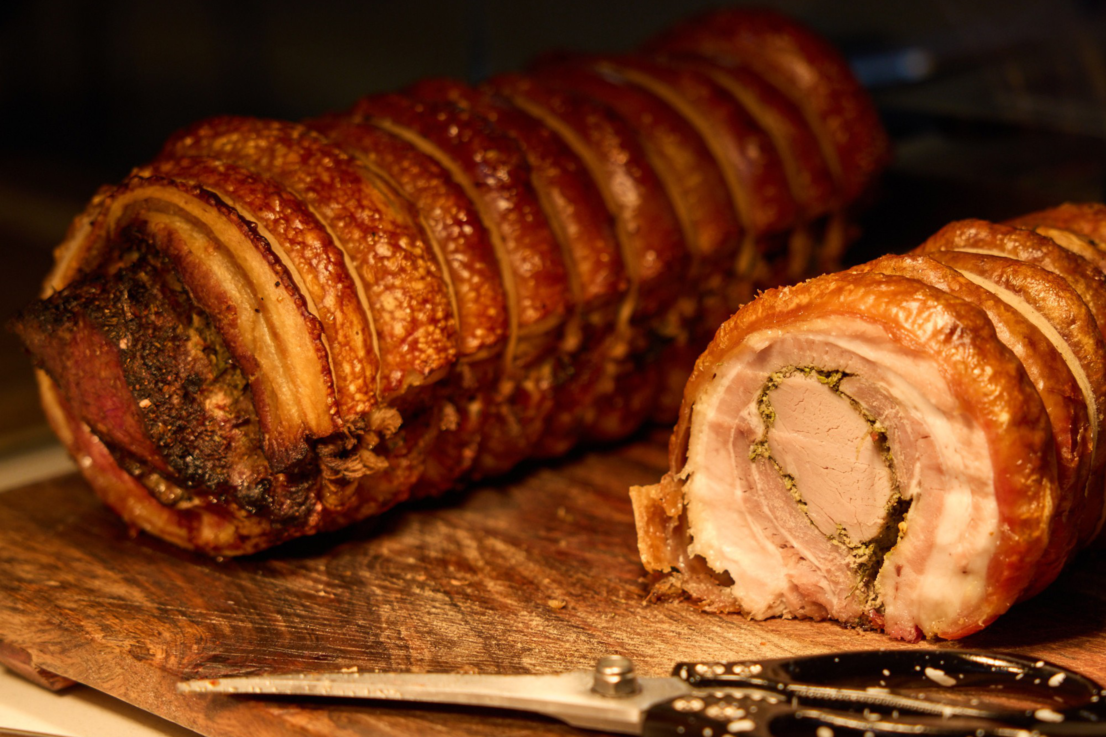 The image shows a rolled and roasted pork on a wooden cutting board. One end is sliced to reveal the inner layers, with kitchen shears placed nearby.