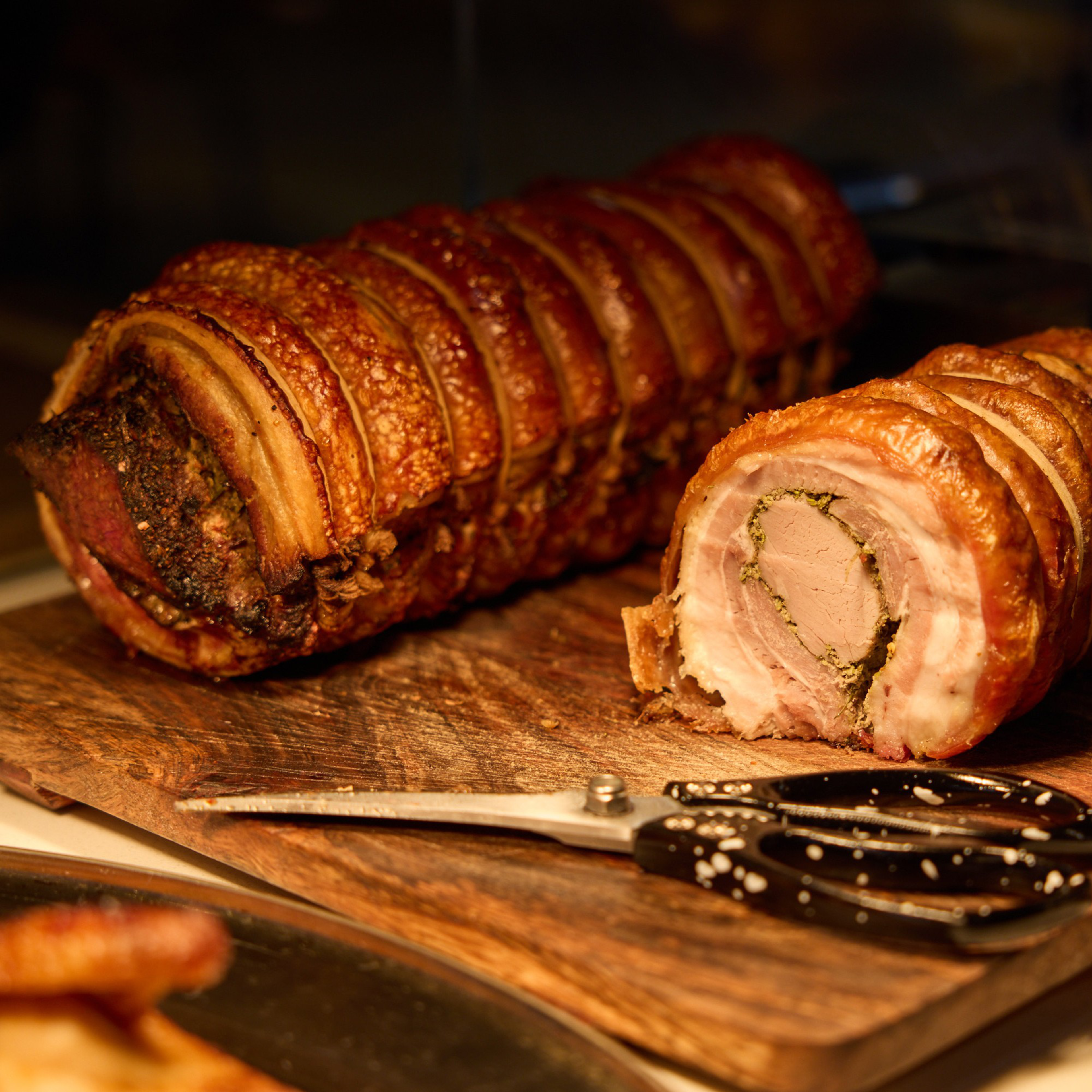 The image shows a rolled and roasted pork on a wooden cutting board. One end is sliced to reveal the inner layers, with kitchen shears placed nearby.
