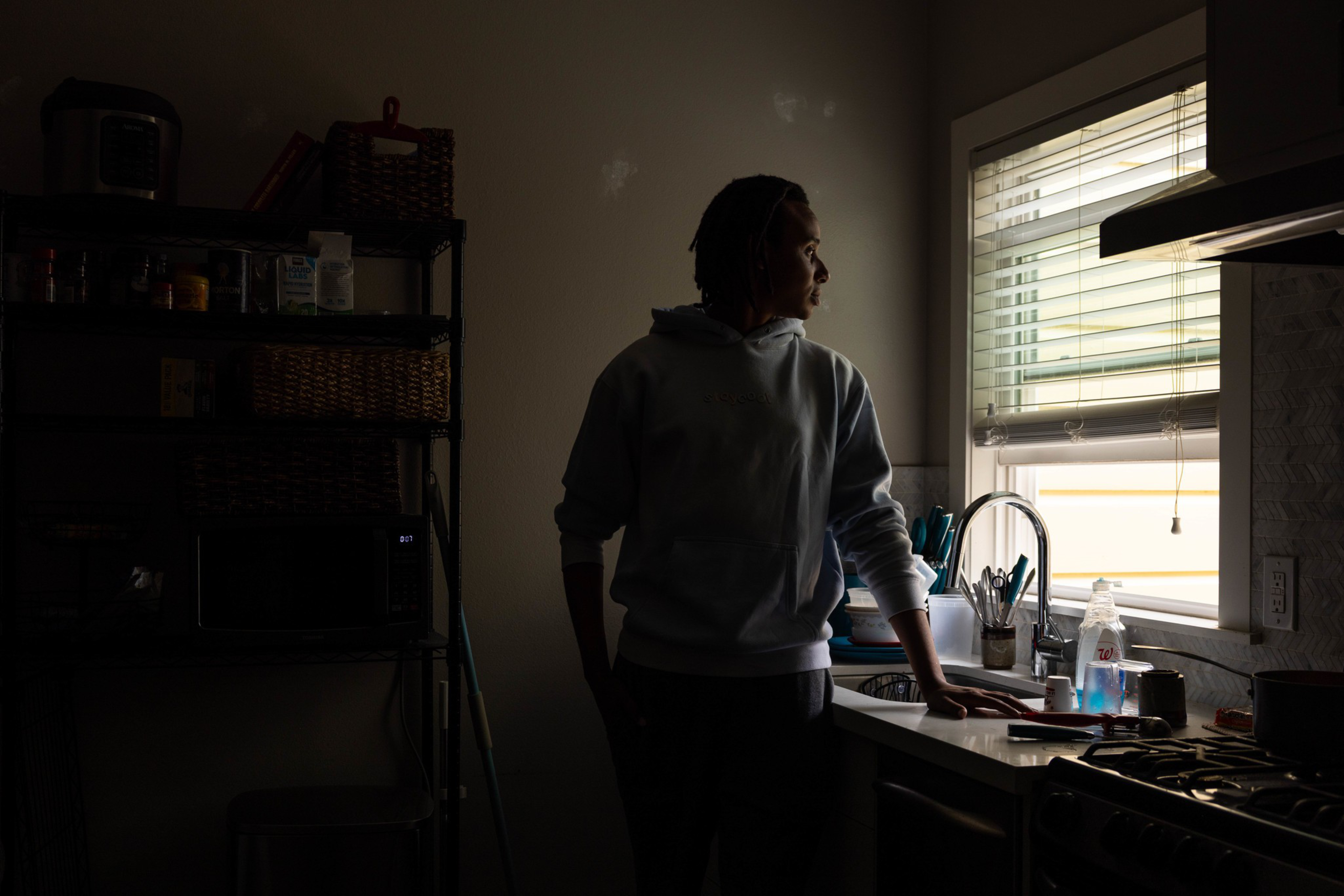 A person in a hoodie stands quietly by a kitchen sink, looking out a window with blinds, with soft daylight illuminating their face.