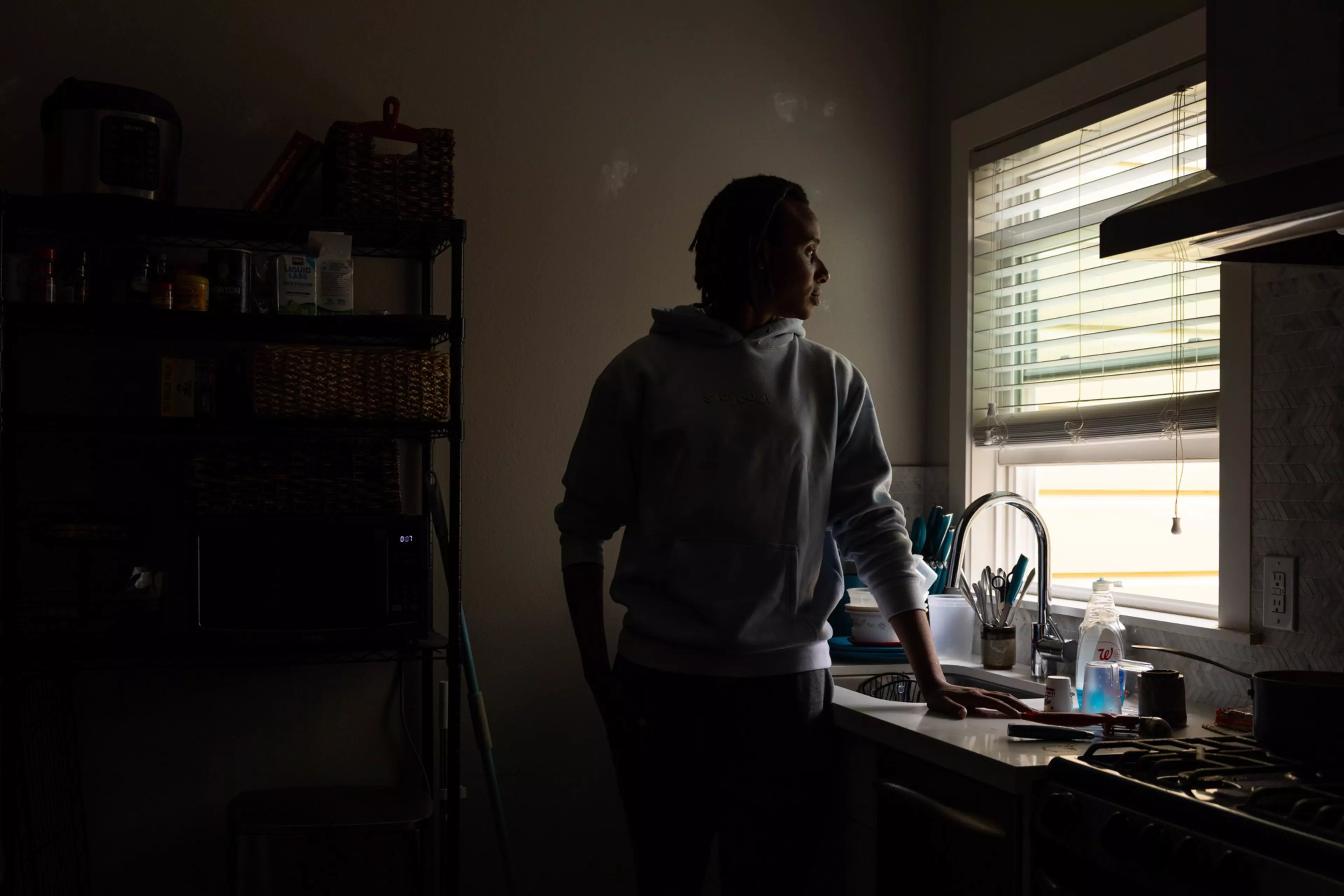 A person in a hoodie stands quietly by a kitchen sink, looking out a window with blinds, with soft daylight illuminating their face.
