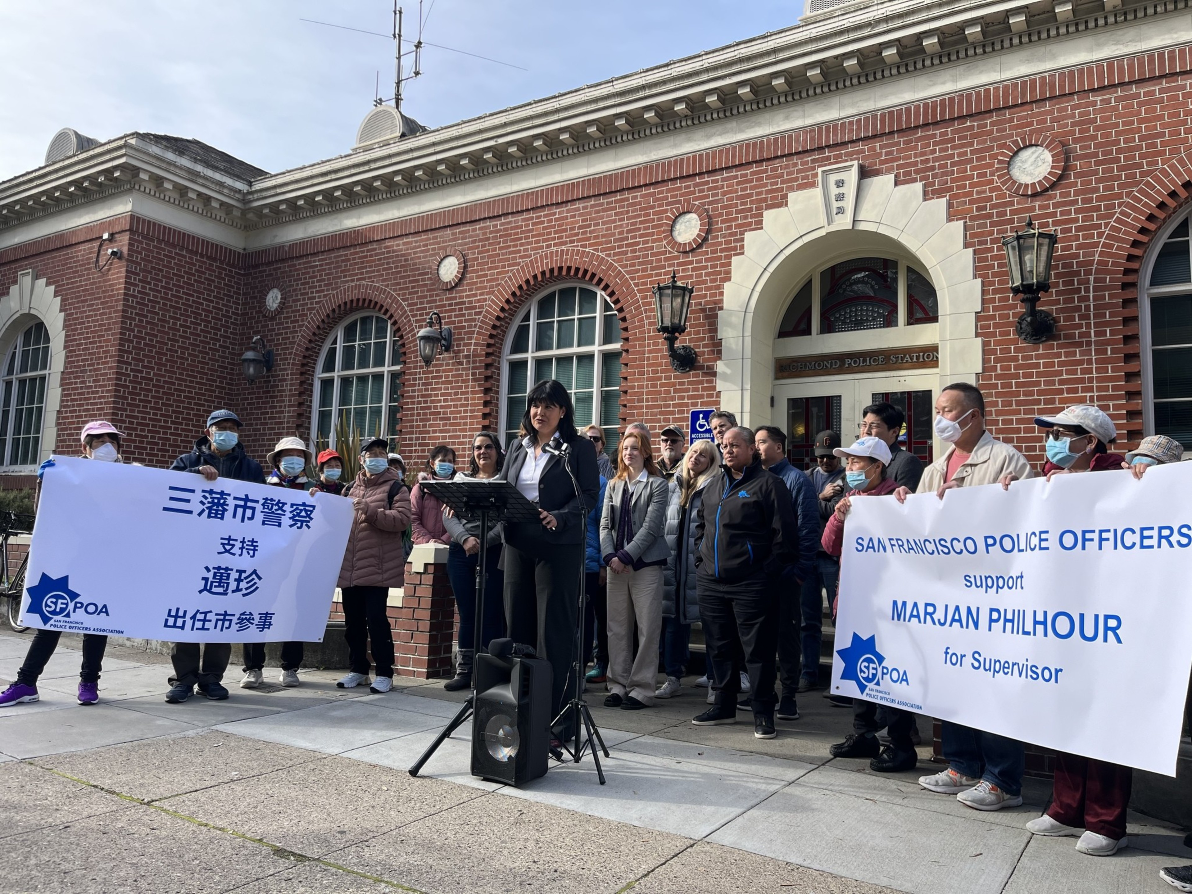 A group is gathered outside a brick building; some hold a banner, and a woman stands at a podium speaking.