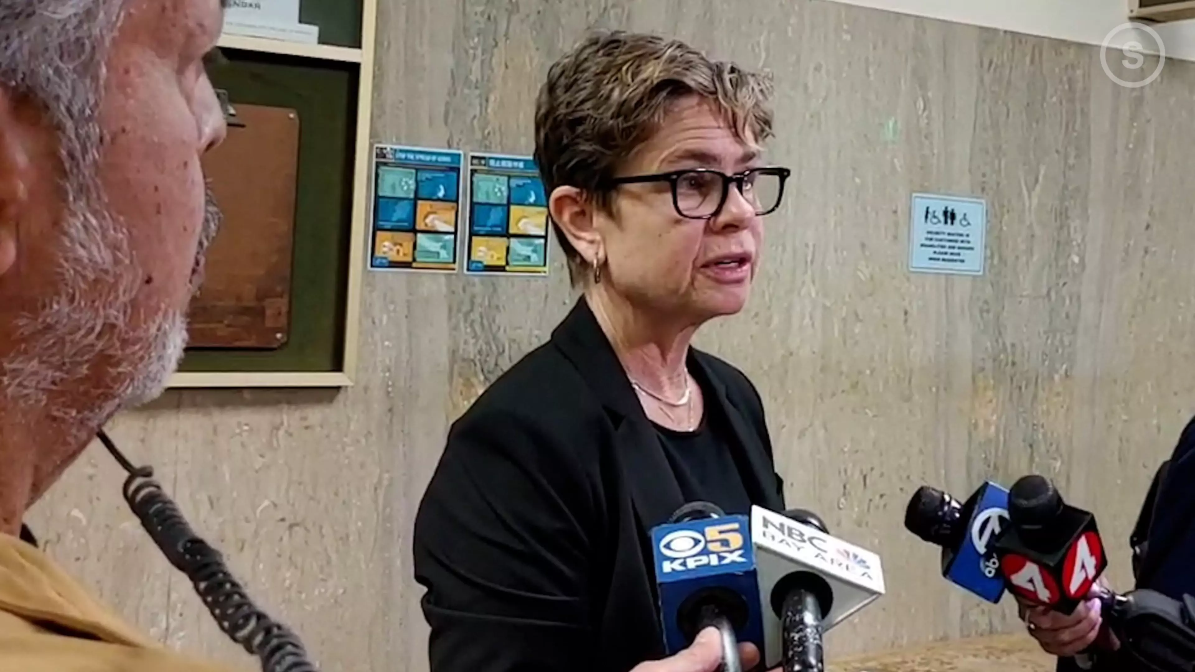 A woman speaks into microphones held by reporters, with onlookers and a man's profile visible.