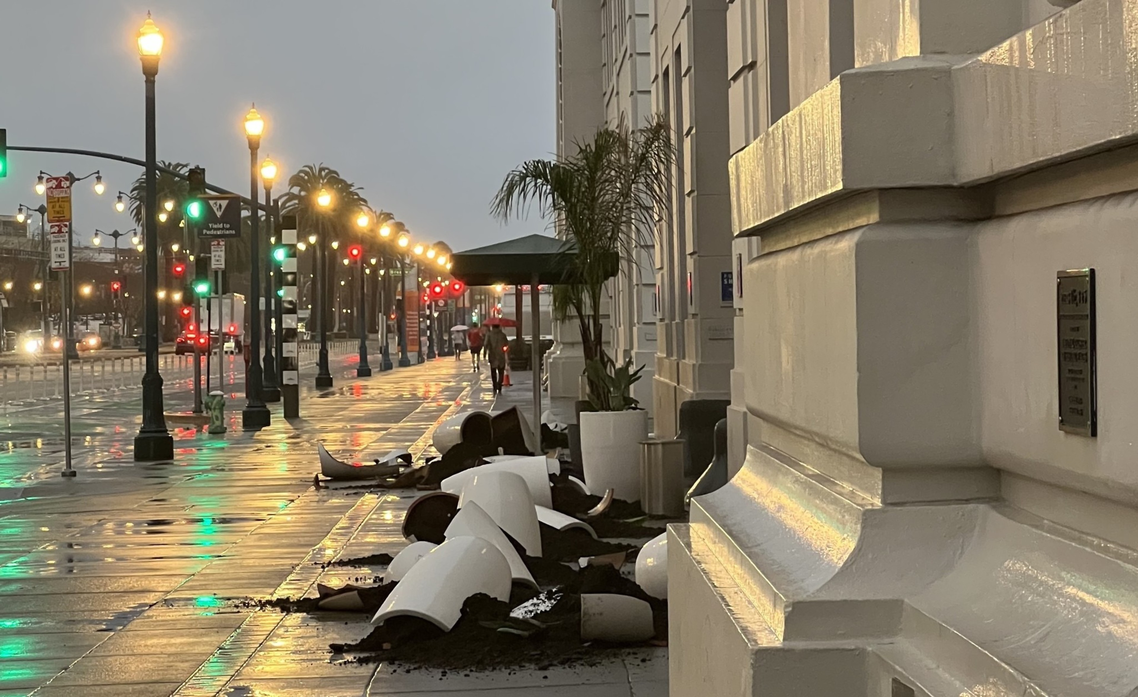 A wet street at dusk with rows of street lamps, toppled planters, and a distant figure with an umbrella.