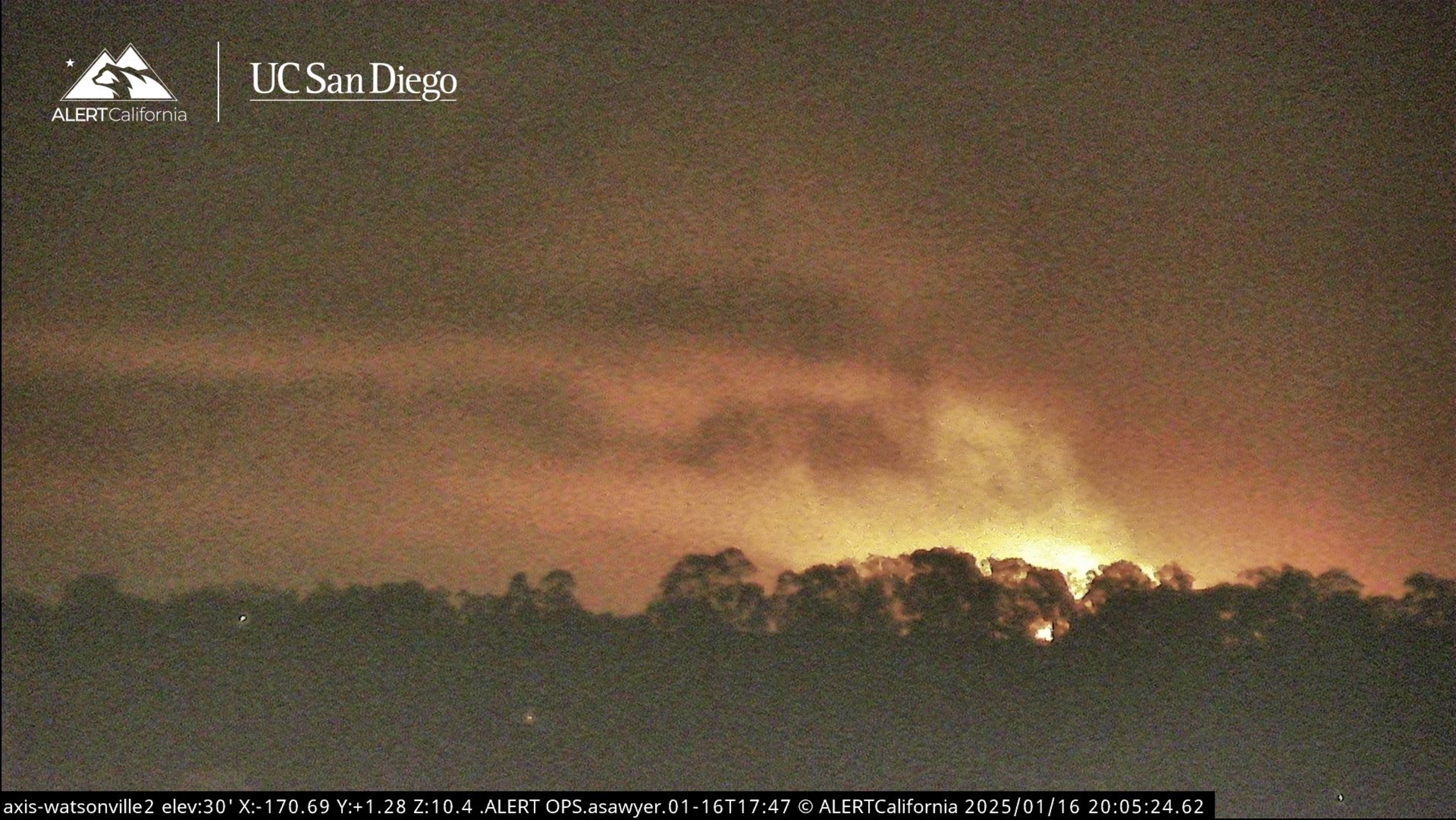 The image shows a wildfire with bright flames and smoke rising above a silhouette of trees under a night sky. It is branded with UC San Diego and ALERT California.