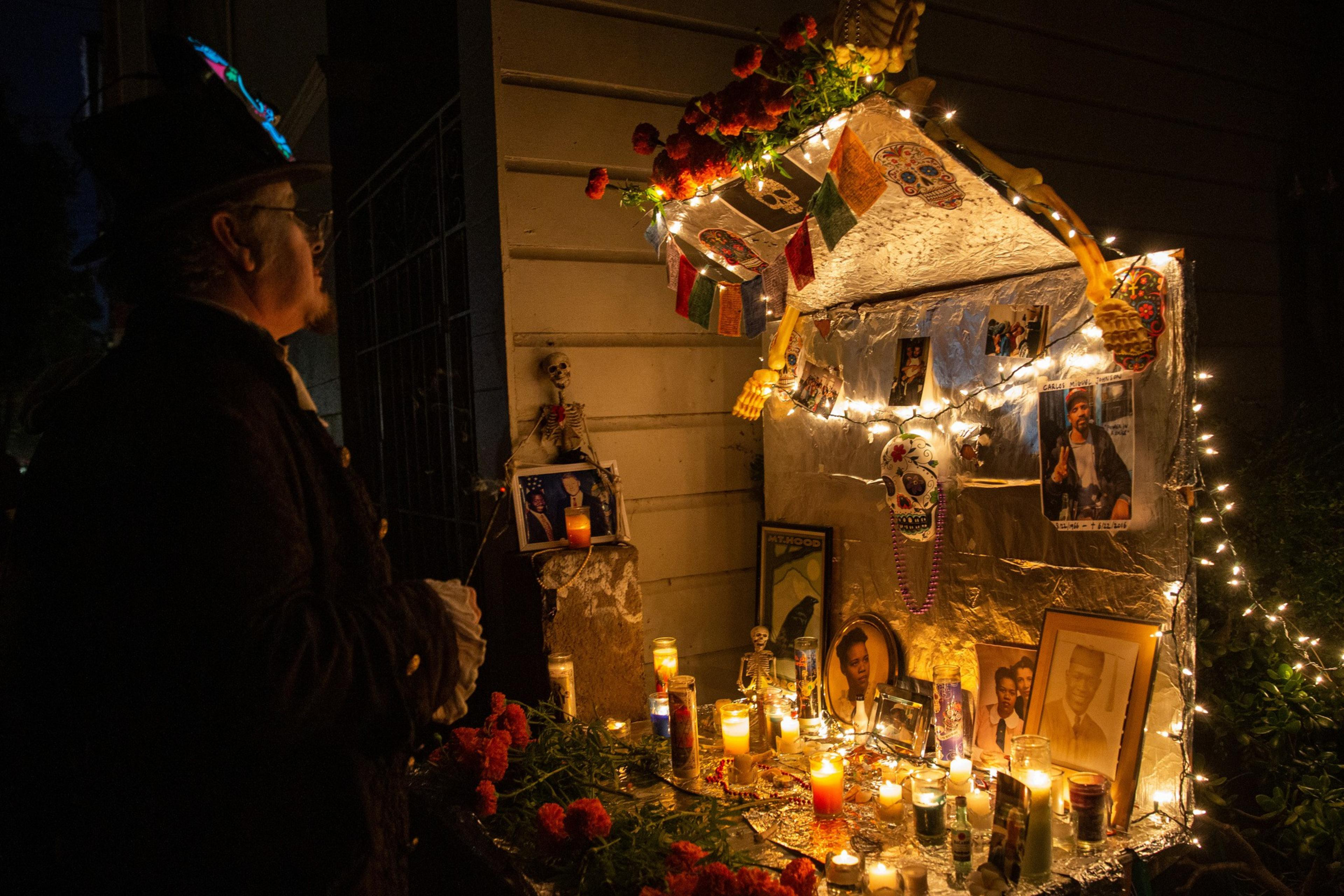 A person wearing a decorated hat and dark clothing stands beside a lit Day of the Dead altar adorned with candles, photos, sugar skulls, flowers, and string lights.