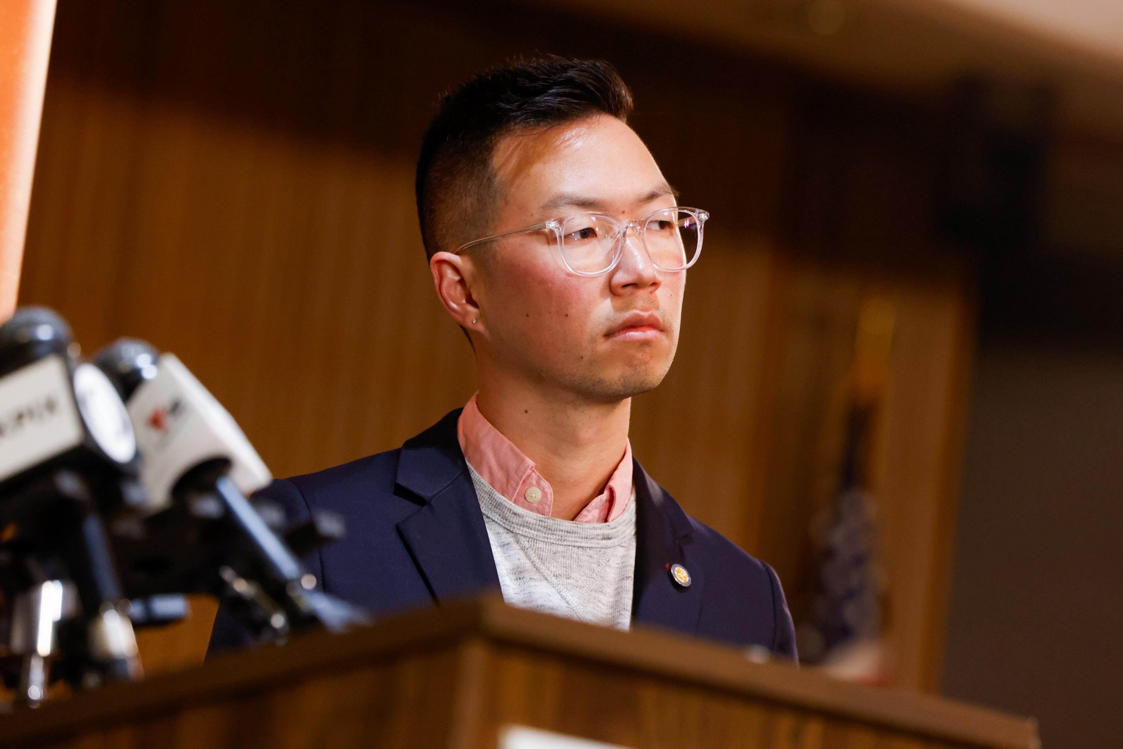 A person with short hair and glasses stands behind a podium with microphones, wearing a suit and a pink shirt, looking serious. An American flag is blurred in the background.