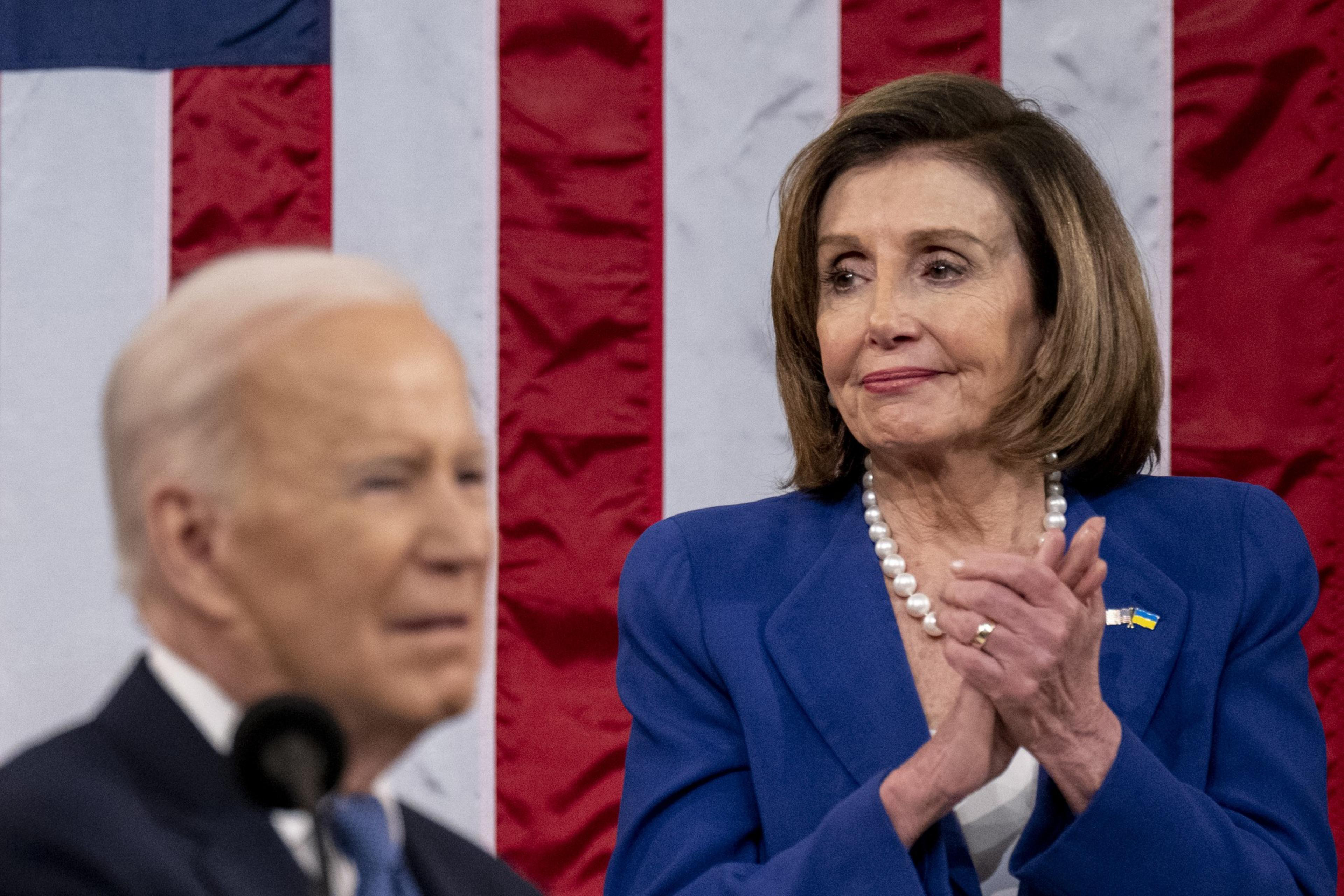 A woman in a blue suit claps with a US flag backdrop. A man in a suit speaks at a podium in the foreground. The scene is formal and patriotic.