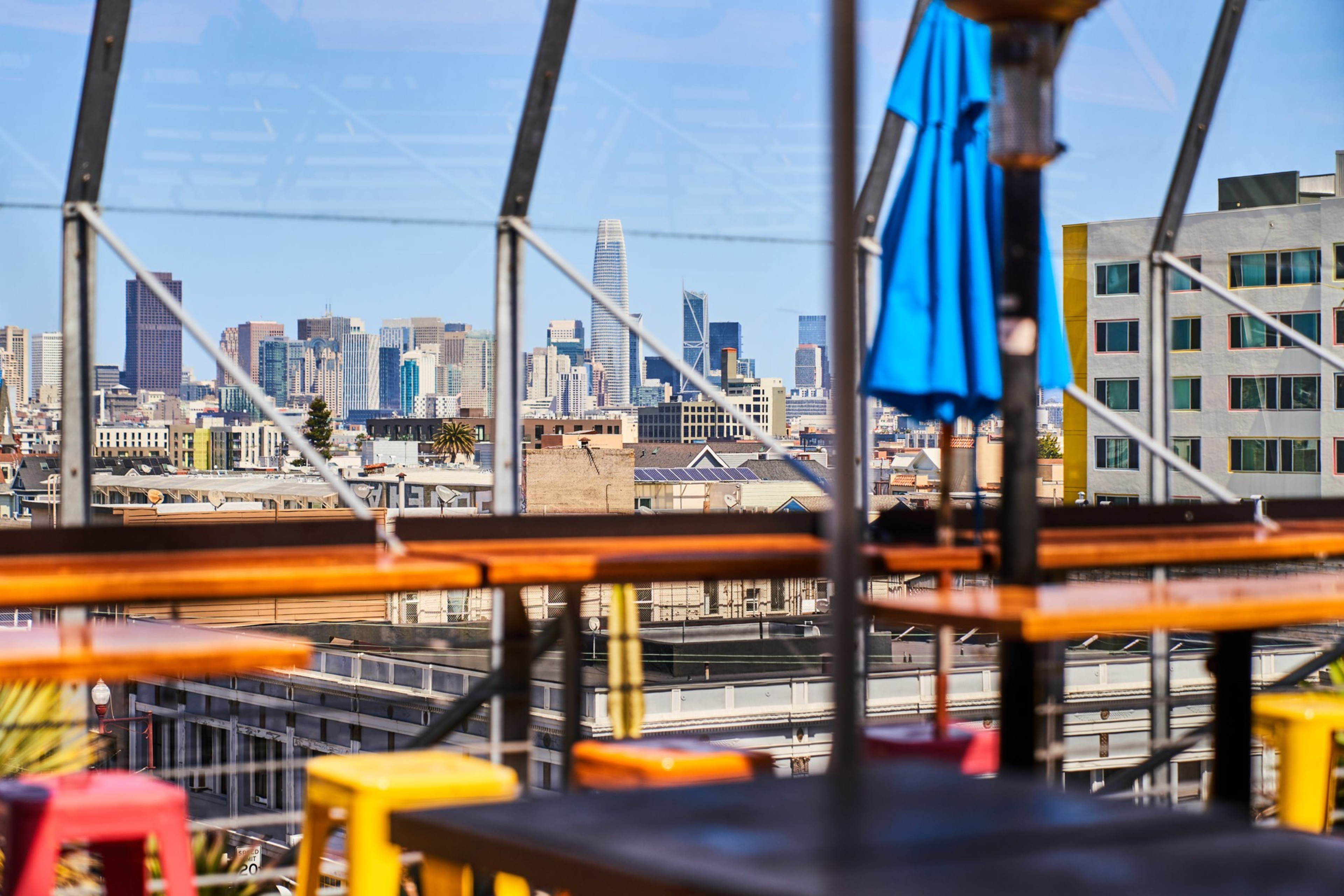 A vibrant rooftop bar offers a view of a city's modern skyline, with colorful stools and tables in the foreground, under a clear blue sky.