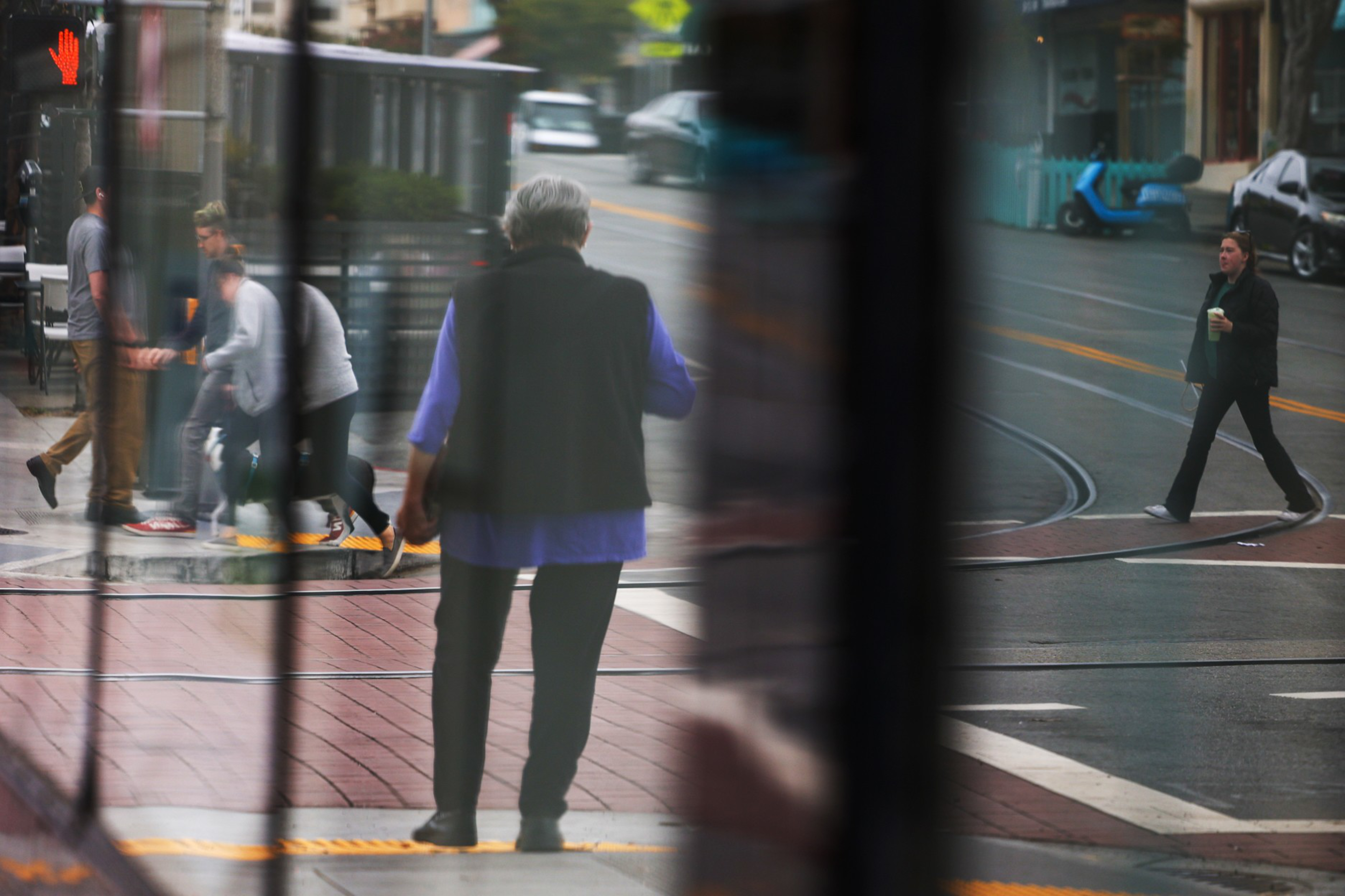 An older person stands still near a tram, while several people walk across the street and another person crosses with a drink.
