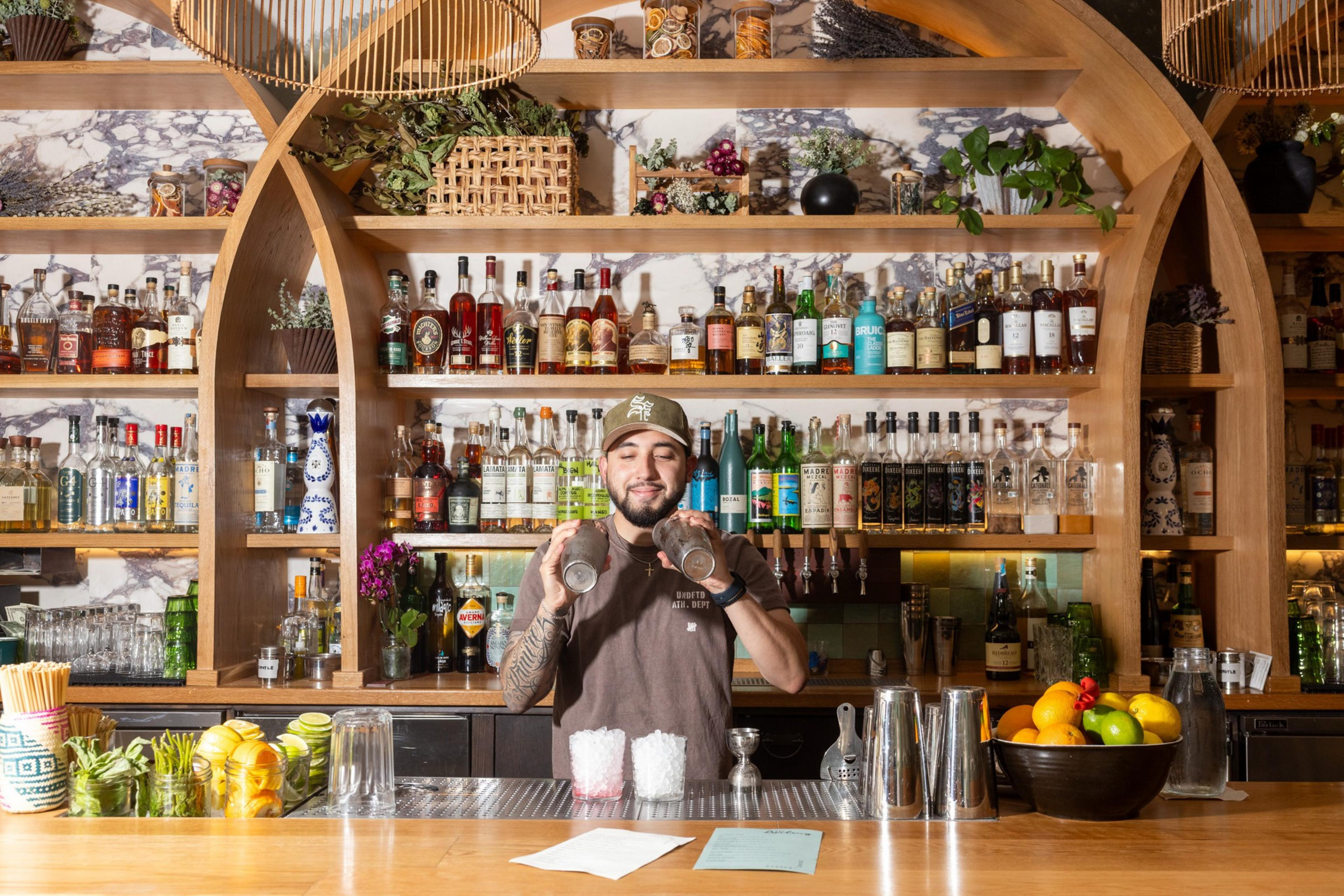 A bartender is smiling while mixing drinks behind a wooden bar. The shelves are stocked with various liquor bottles, and a bowl of fresh fruits sits on the counter.