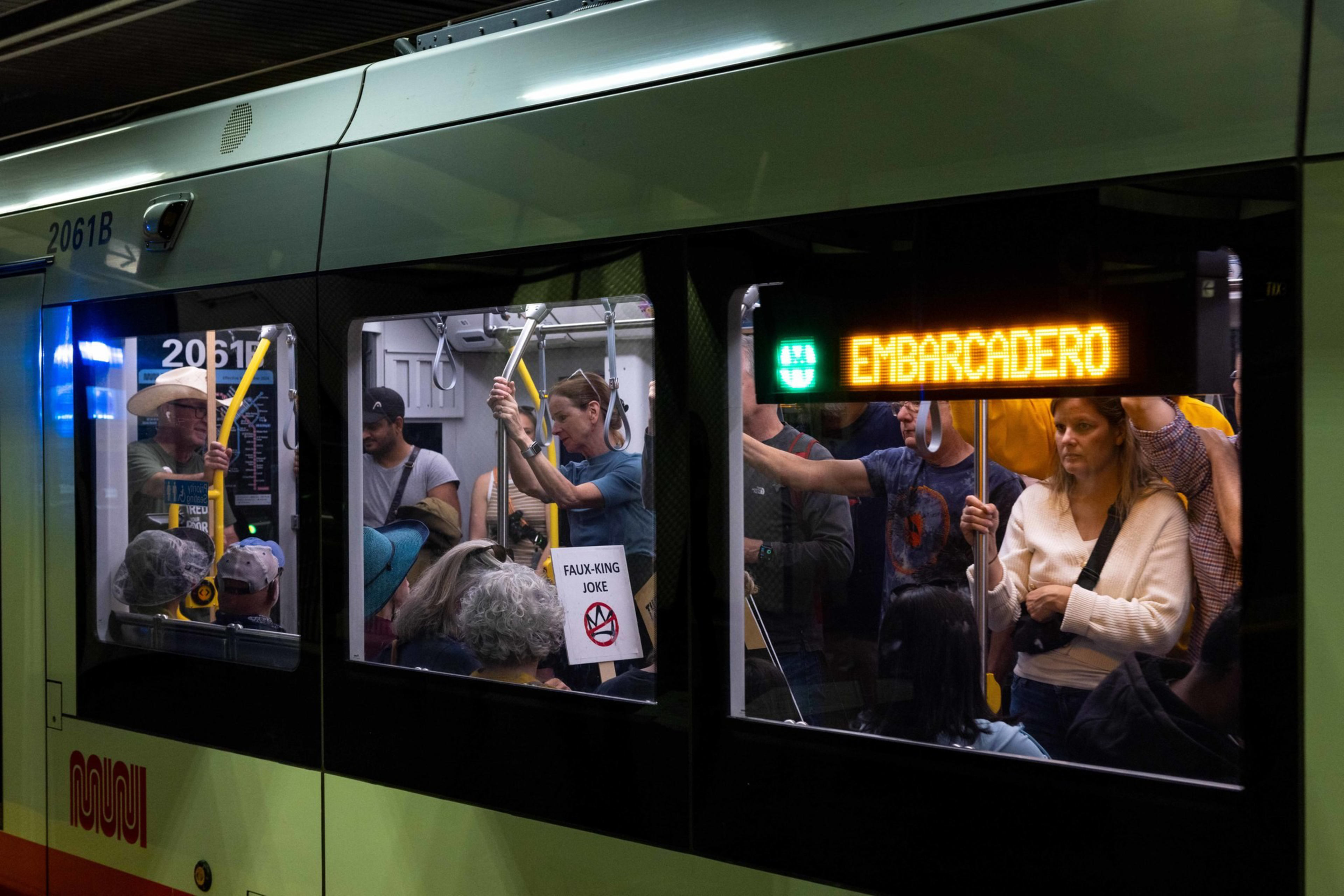 People stand and sit inside a crowded Muni train with a digital sign showing the destination “EMBARCADERO” at night.