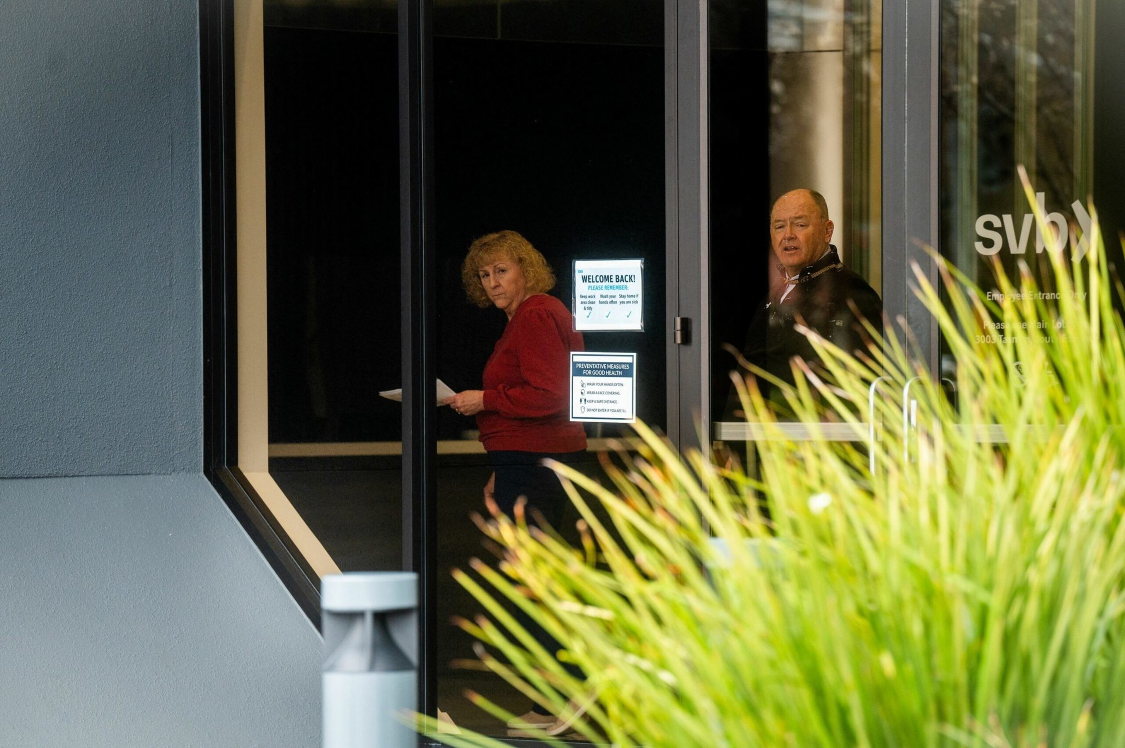 A woman in a red sweater and a man in dark clothing stand inside a glass-entry building, partly obscured by tall green grass outside.