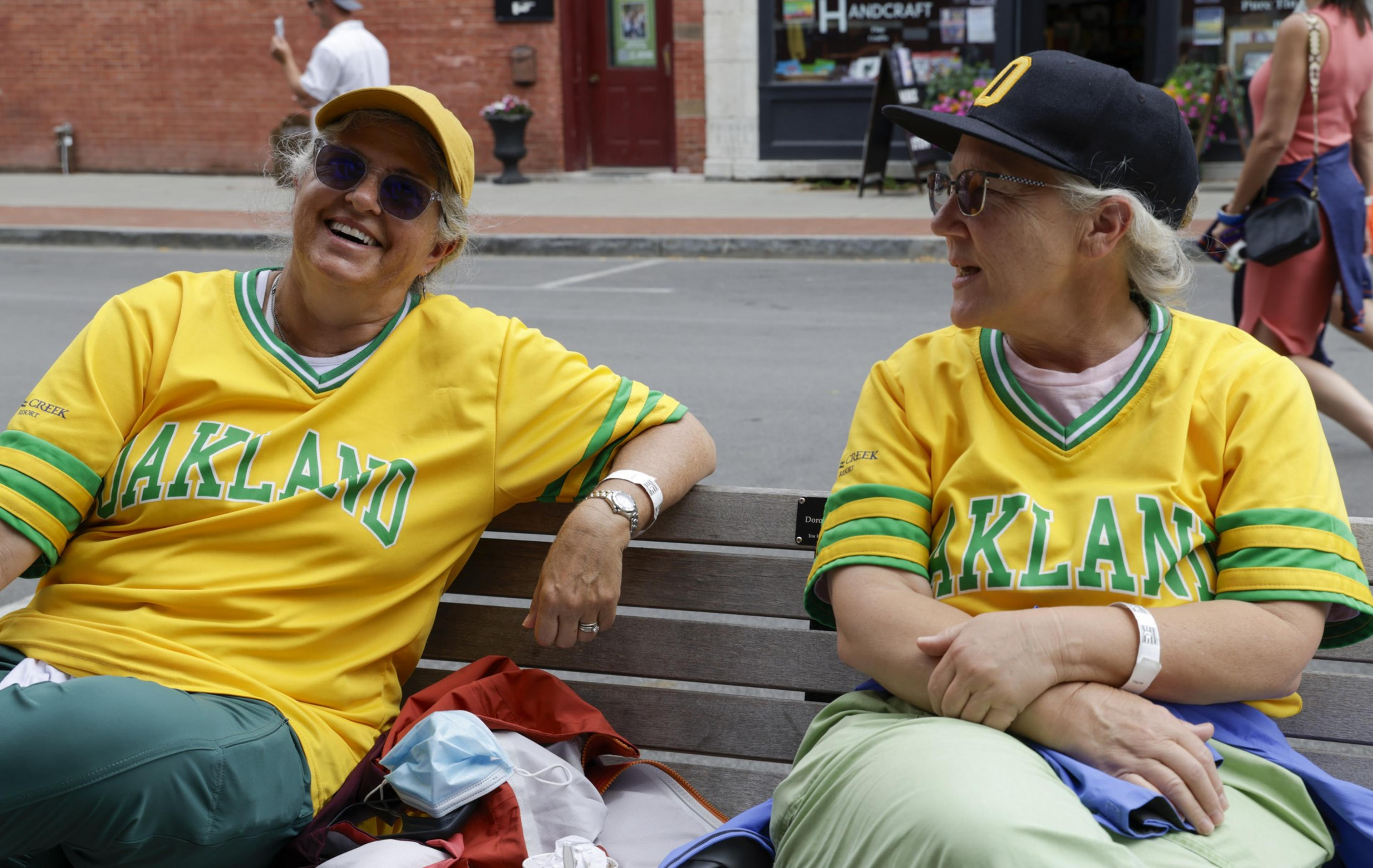 Two people are sitting on a bench wearing matching yellow &quot;Oakland&quot; jerseys and hats. They are smiling and appear to be enjoying a conversation.