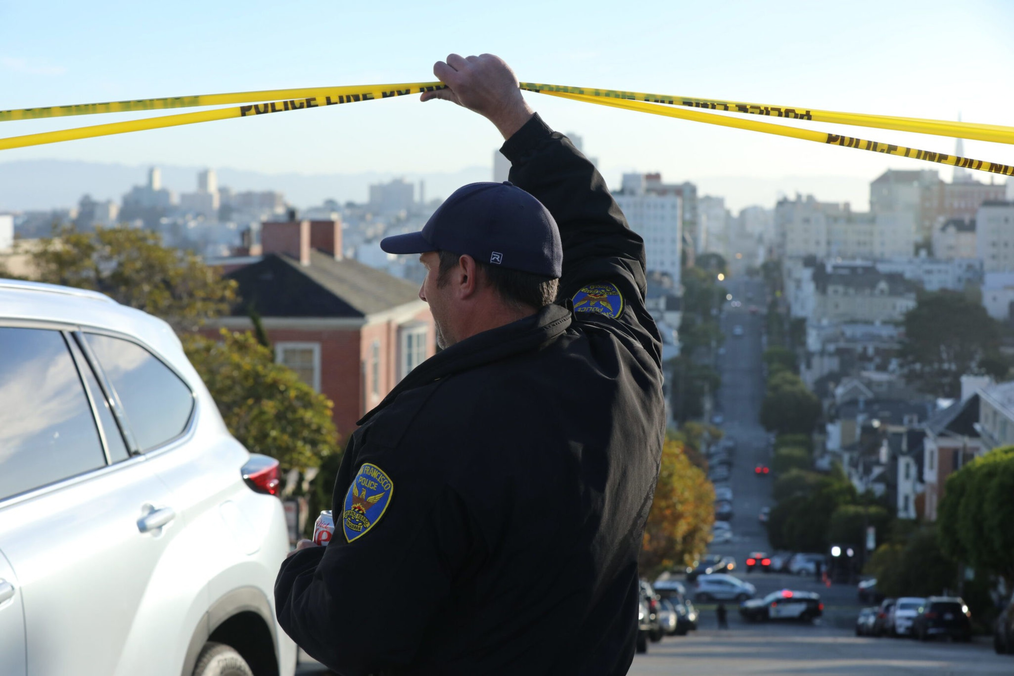 A police officer secures yellow police tape across a street with a cityscape and vehicles in the background during daylight.