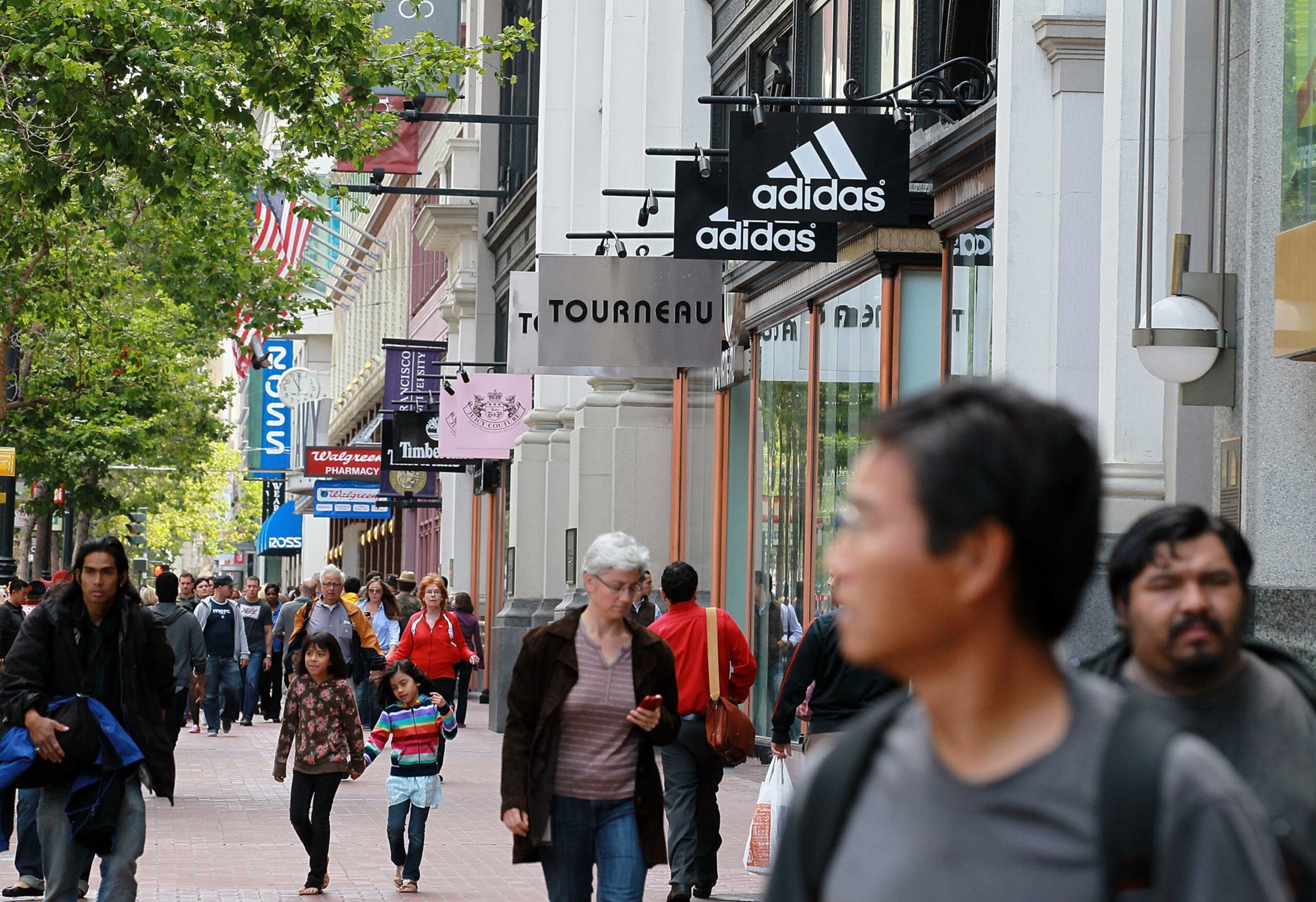 A busy city sidewalk with people walking past storefronts, including signs for Adidas, Tourneau, Walgreens, and Ross. Trees and flags line the street.