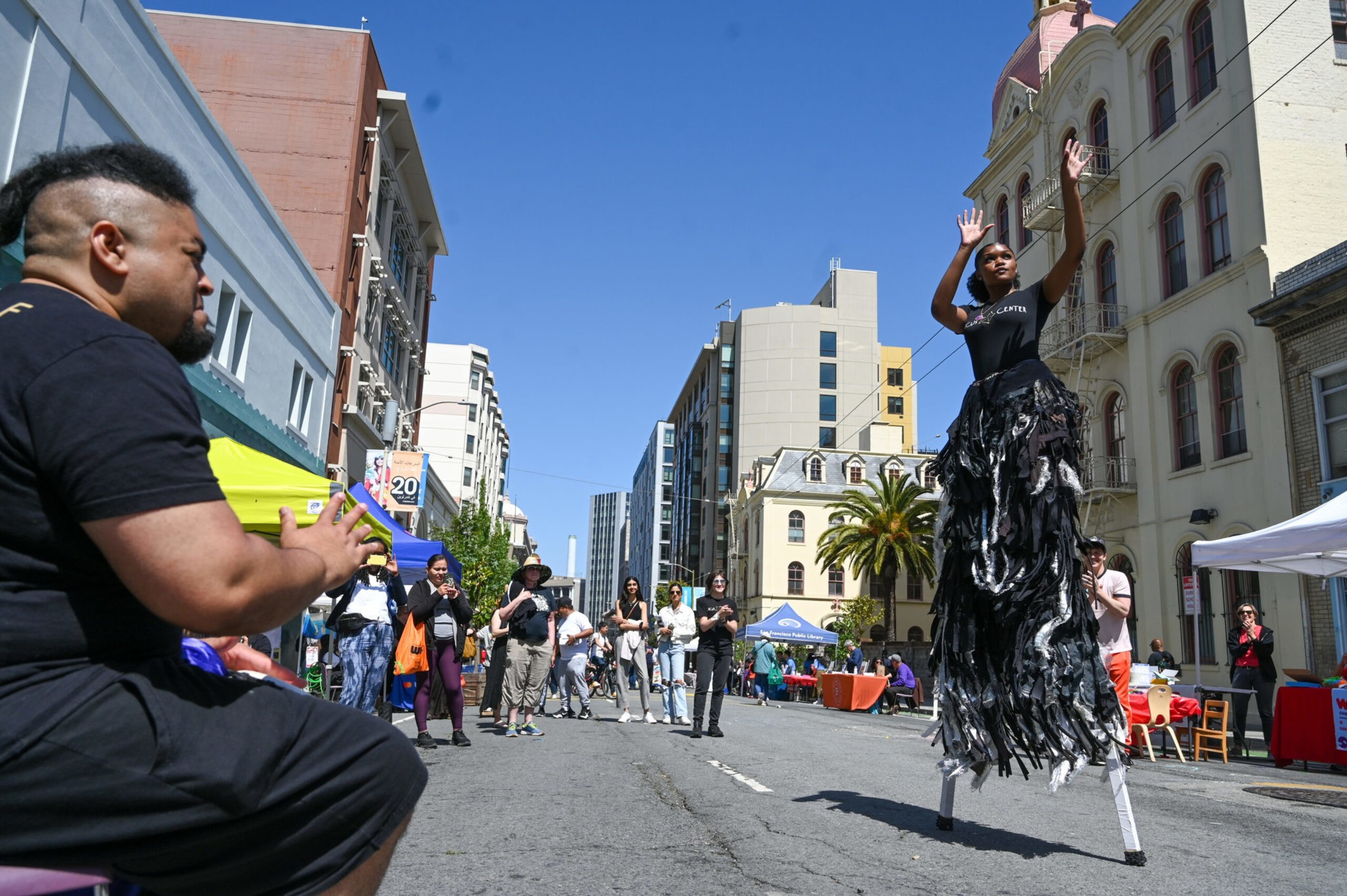 A person on stilts is performing in a street, surrounded by an audience and urban buildings under a clear sky.