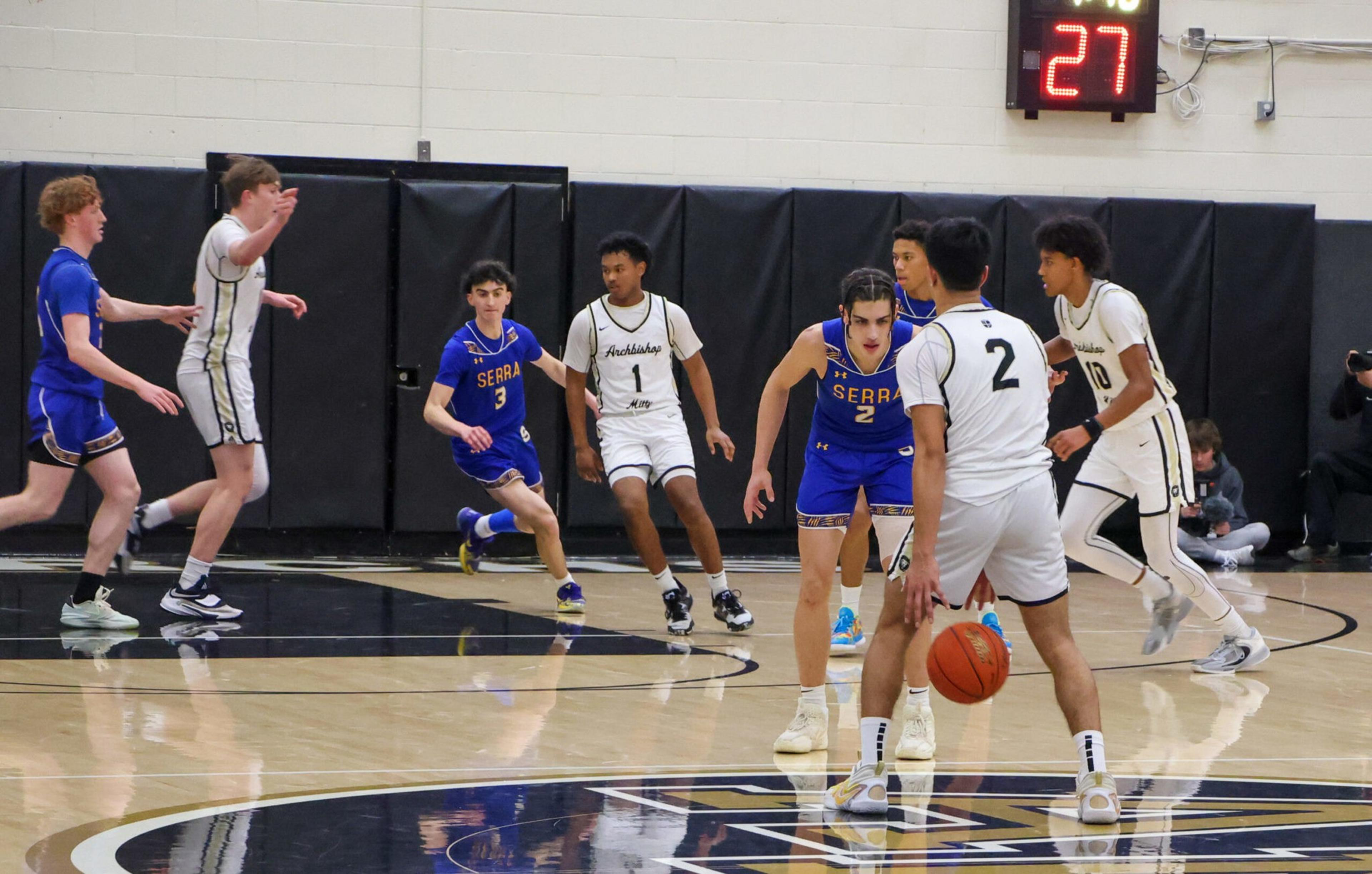 Players in blue and white jerseys are positioned on a basketball court, with one player in white dribbling the ball and a shot clock showing 27 seconds.