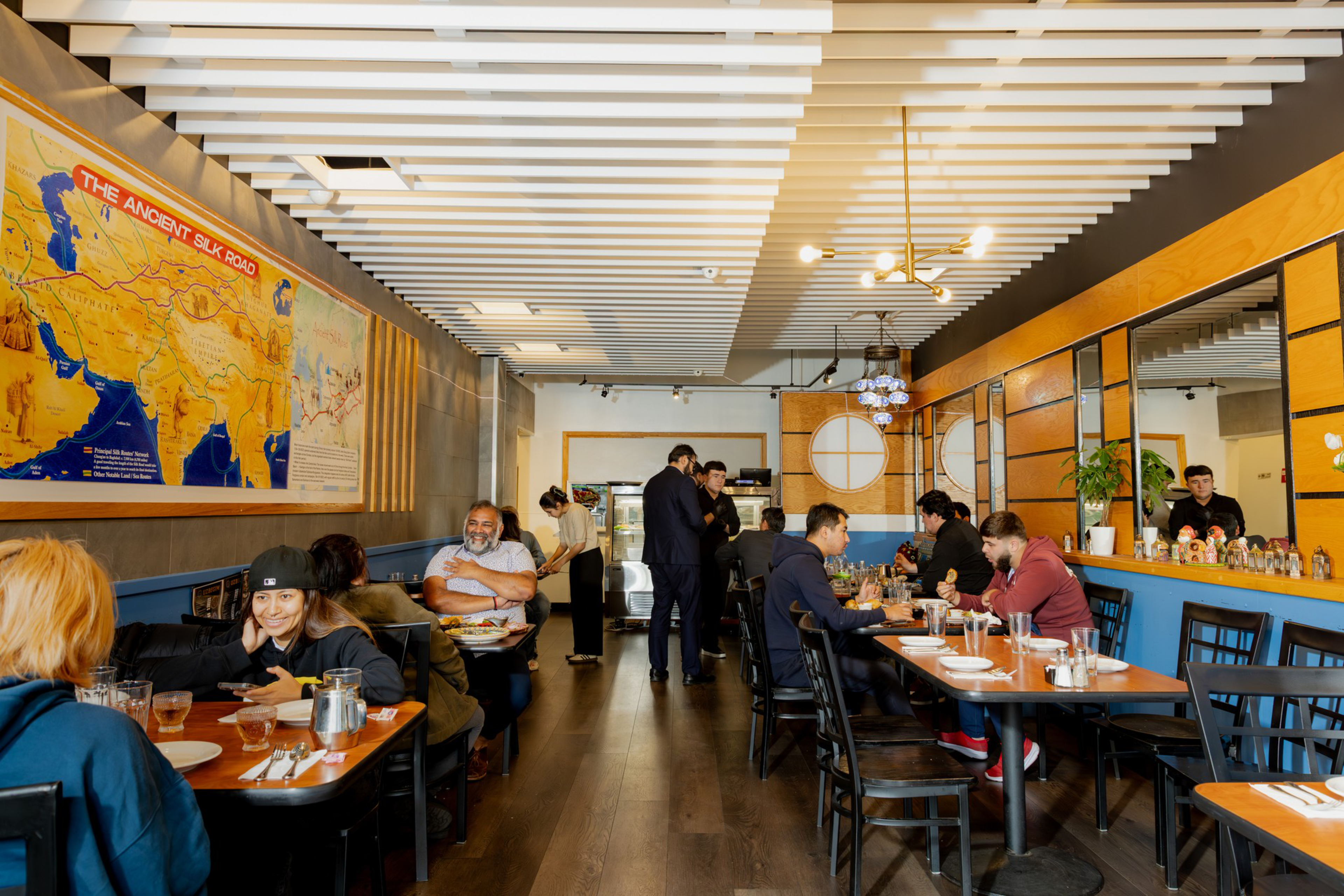 A cozy restaurant with patrons dining and chatting, wooden tables, a large map of the Ancient Silk Road on the wall, and a server attending guests.