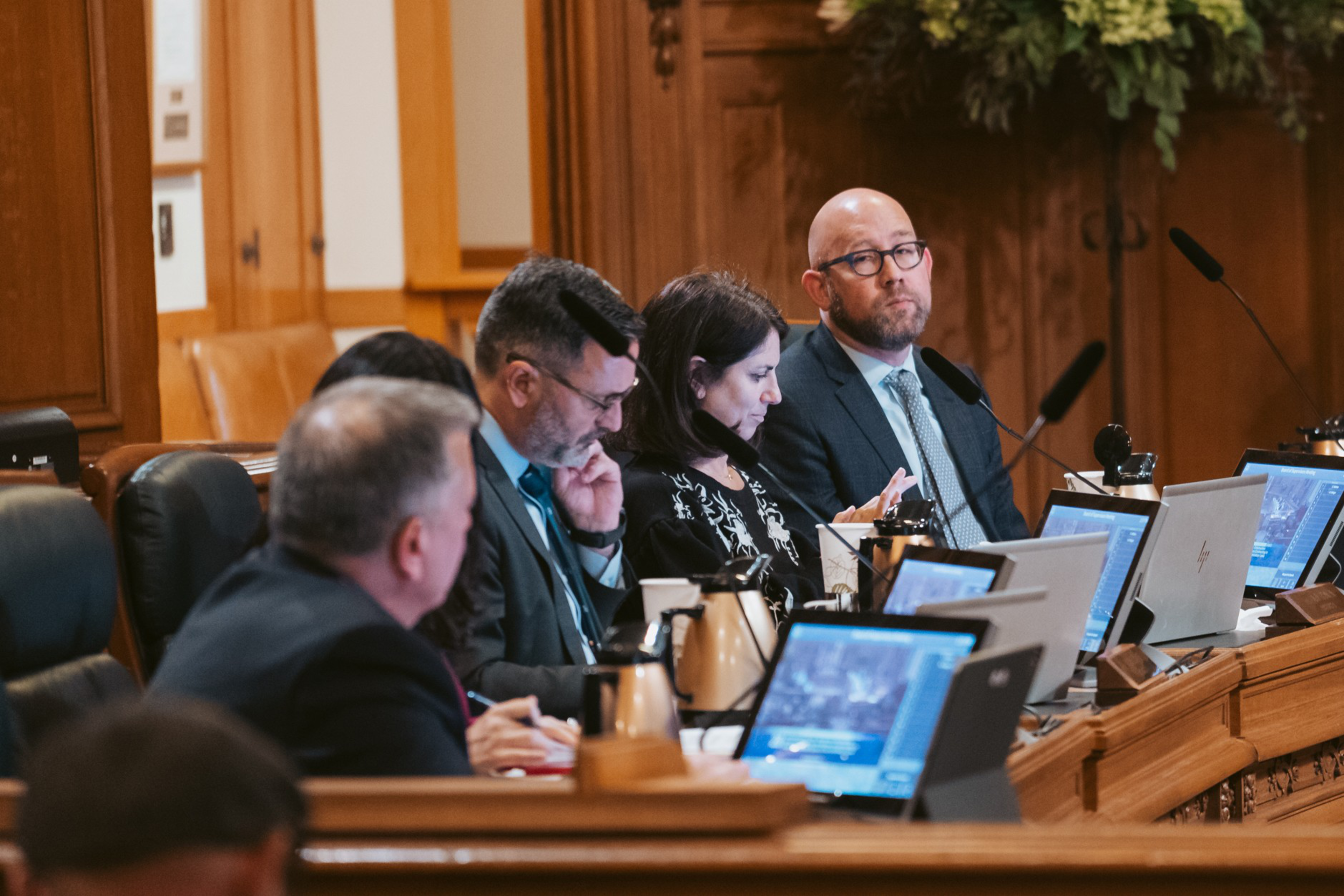 Five people sit at a wooden panel desk with laptops and microphones, engaged in a formal meeting or discussion in a wood-paneled room.