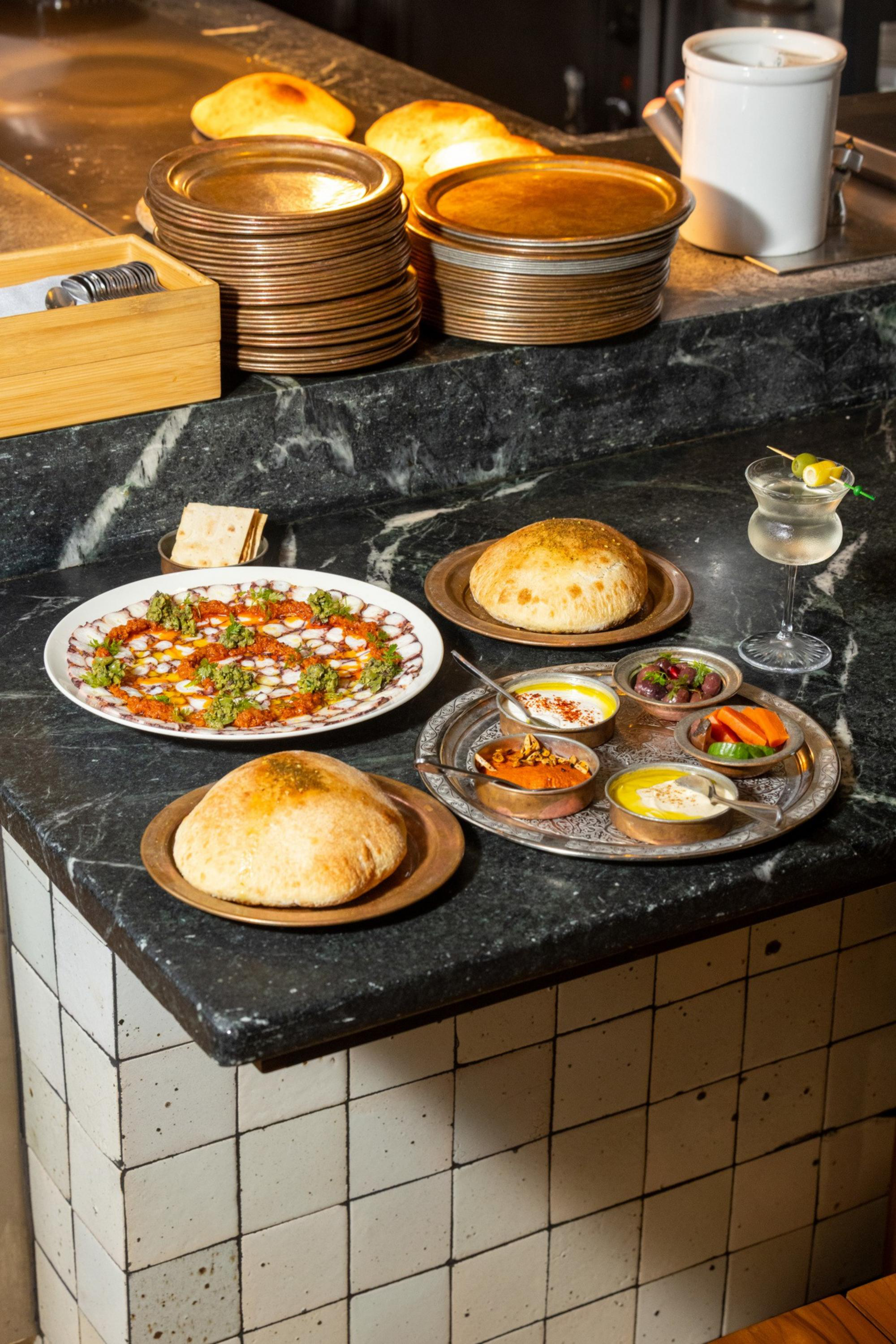 Two plates with round bread, a plate of sliced food garnished with greens, a tray with dips and vegetables, a glass with a skewer, and stacked wooden plates are on a dark counter.