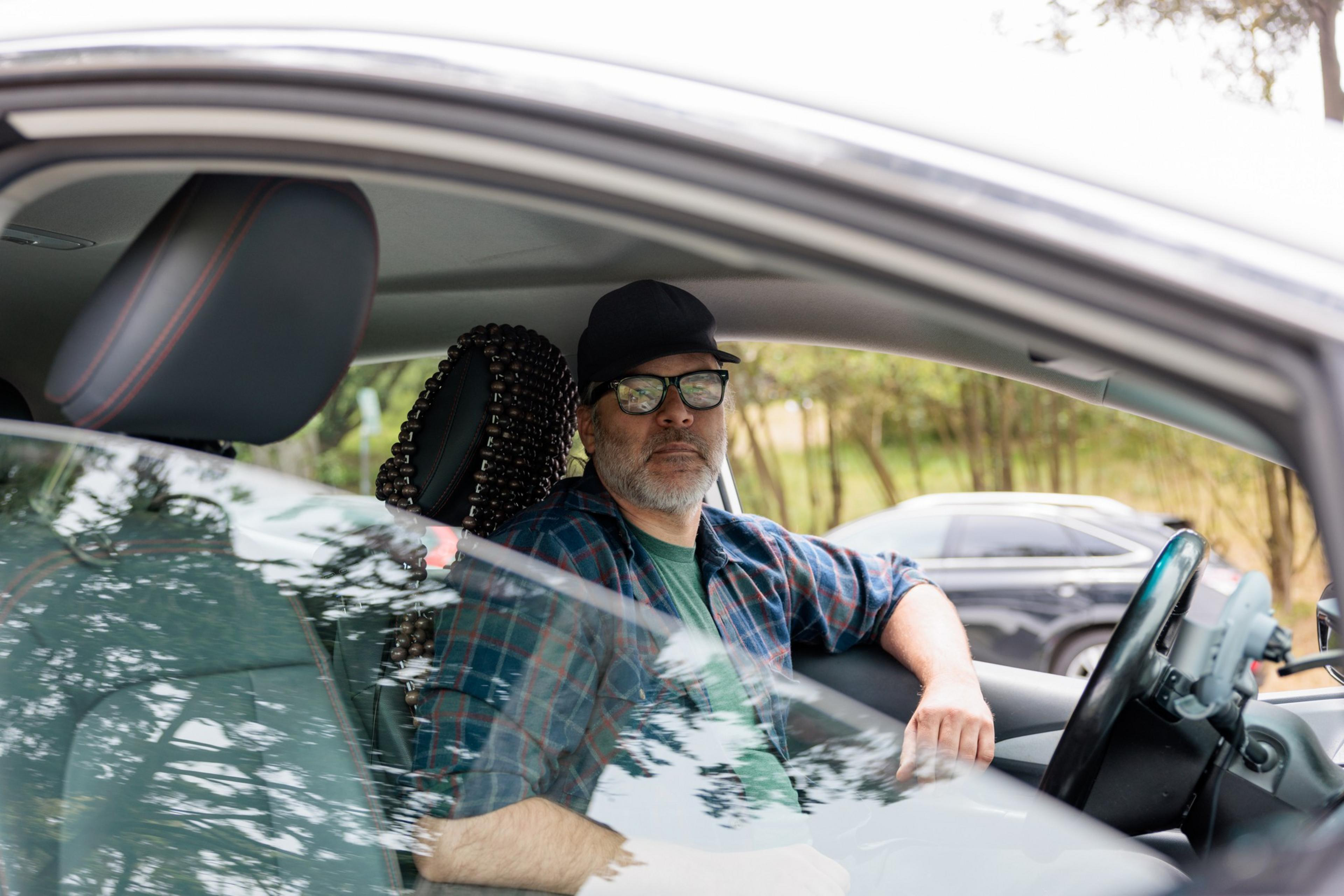 A man wearing a black cap and glasses sits in the driver's seat of a car with a beaded seat cover, looking out the window.