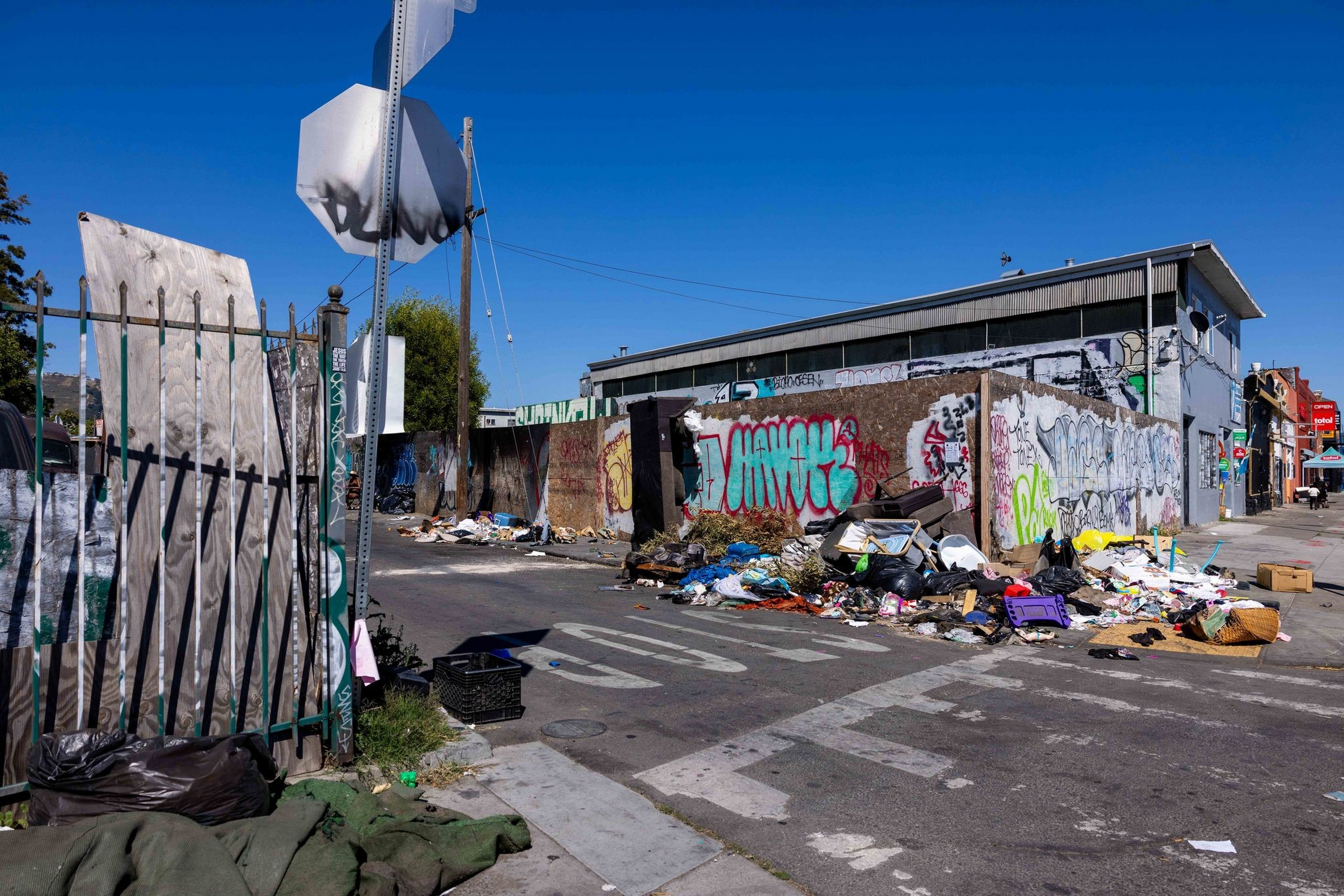 A street corner littered with trash piled against graffiti-covered walls under a clear blue sky.