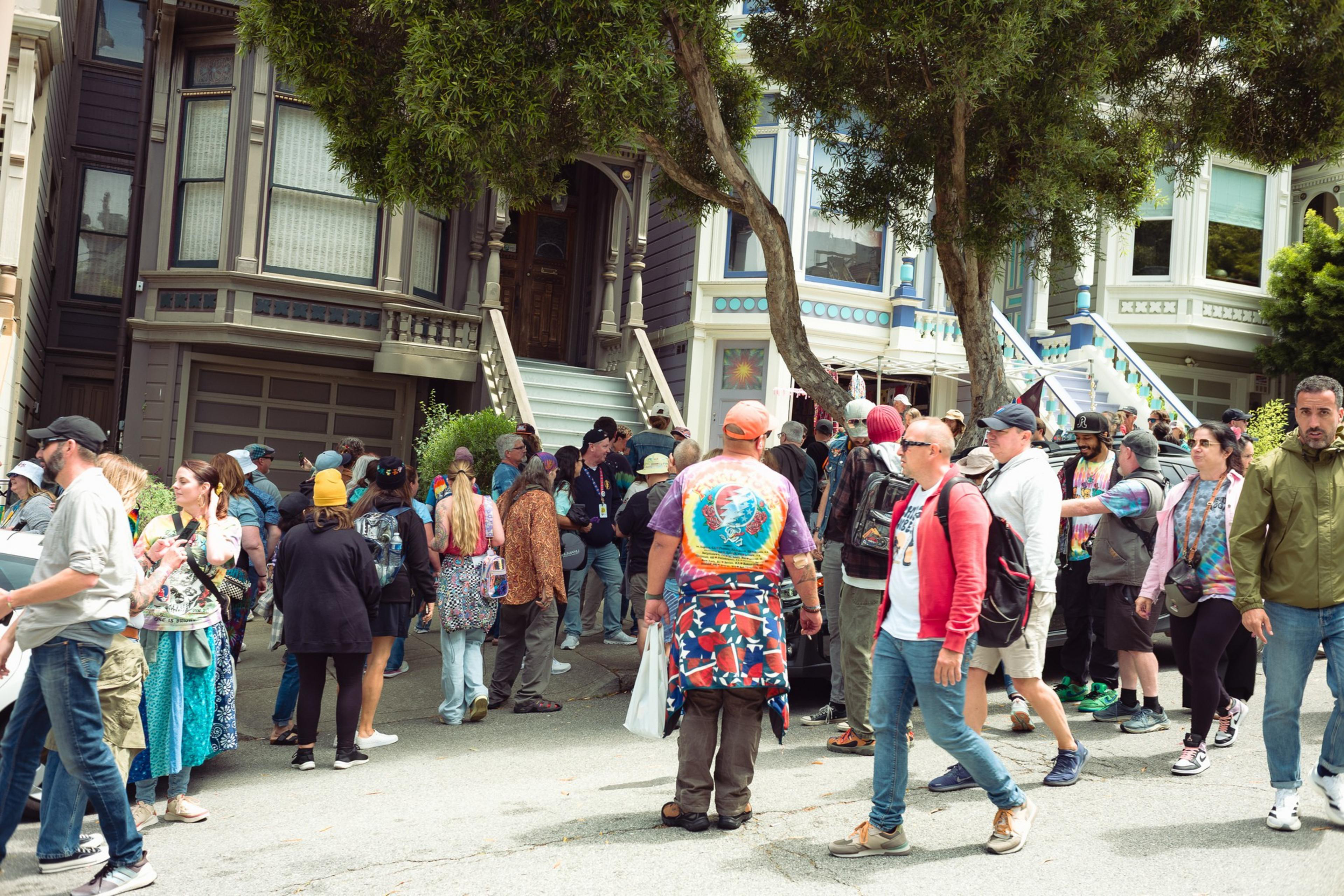 A diverse group of people gathers and walks on a city street lined with colorful Victorian-style houses and trees.
