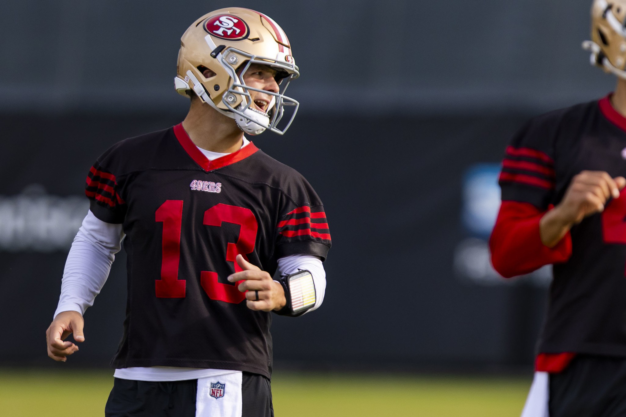 A football player wearing a San Francisco 49ers helmet and black jersey with number 13 gestures while looking to his left.