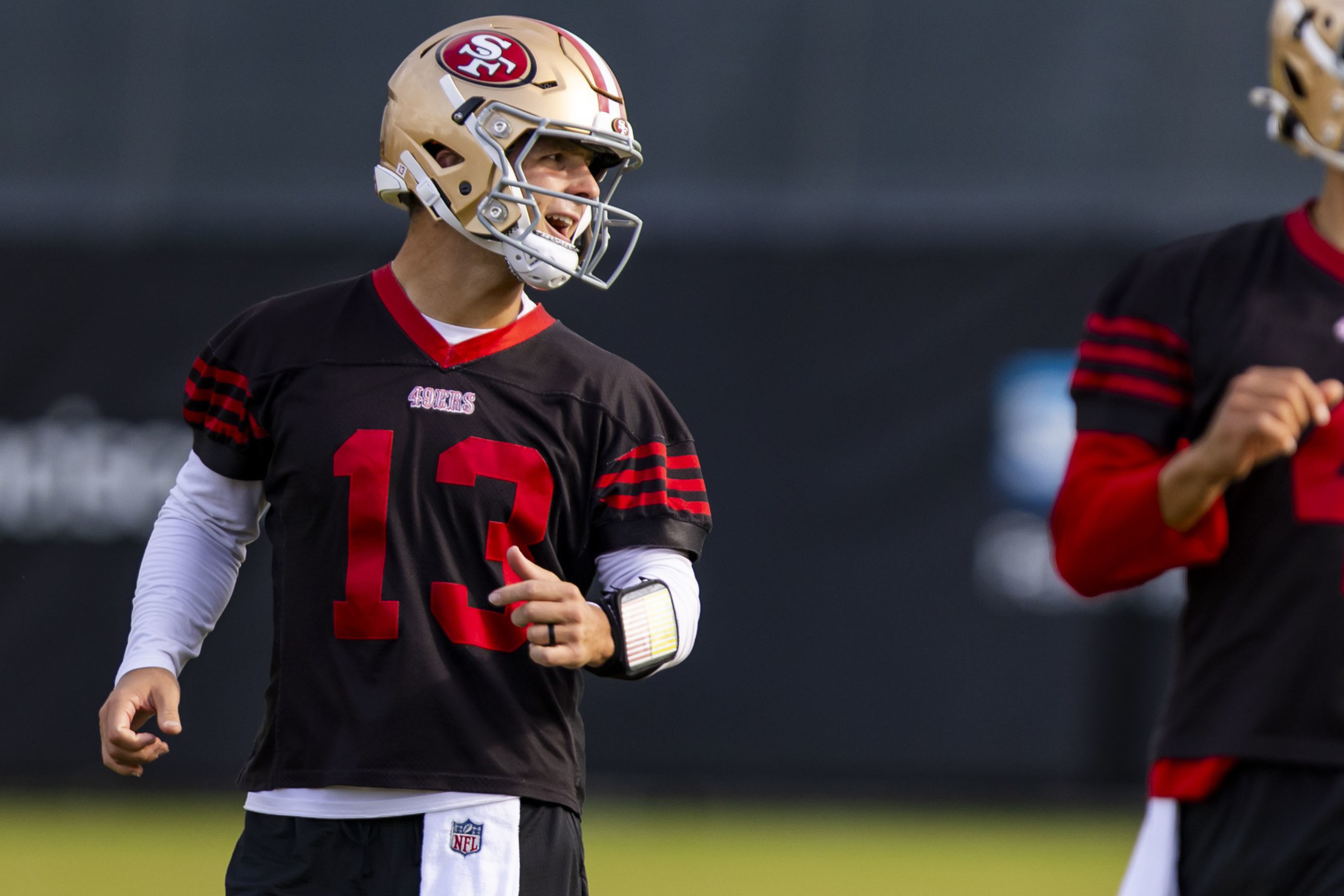 A football player wearing a San Francisco 49ers helmet and black jersey with number 13 gestures while looking to his left.