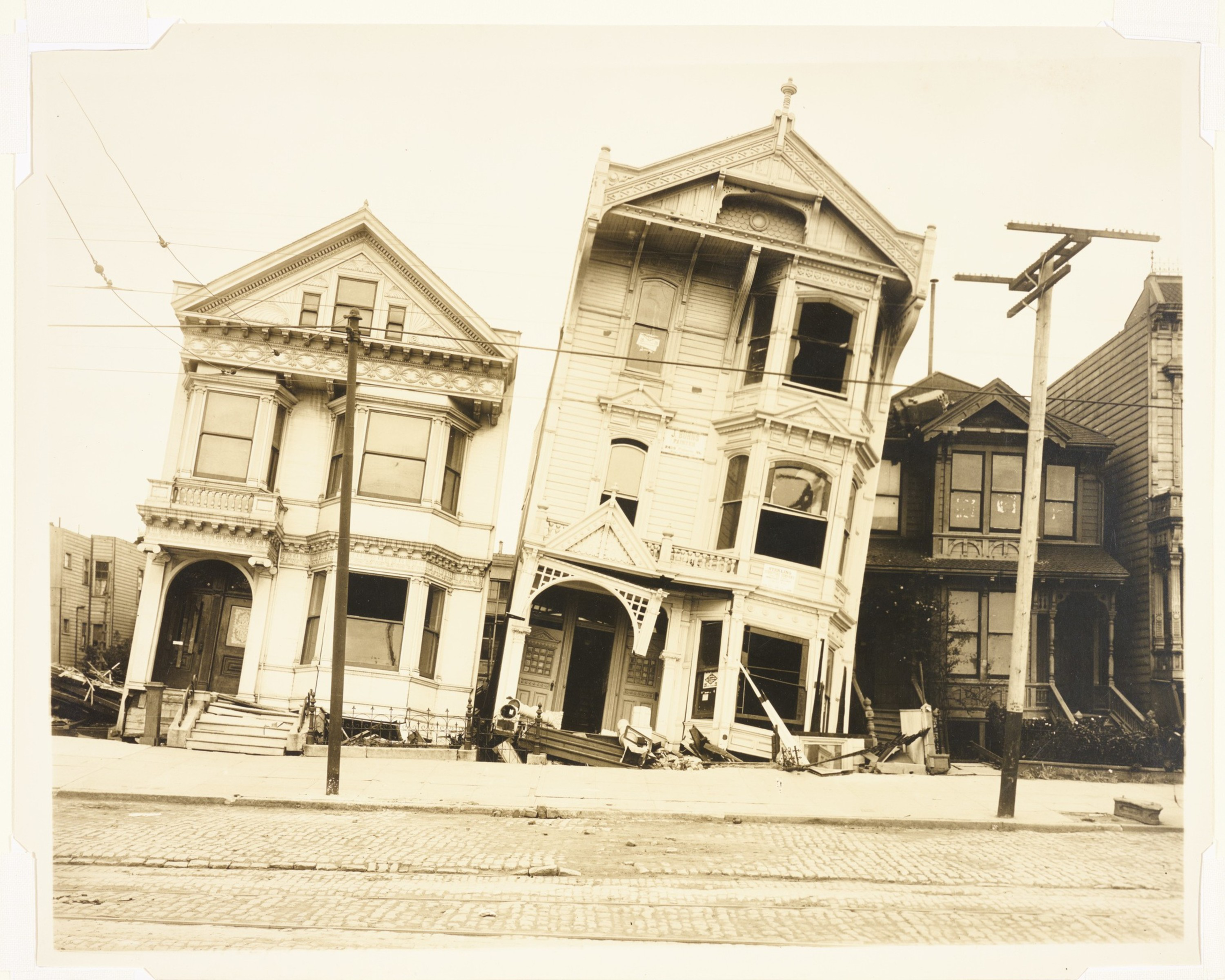 Three old Victorian-style houses appear heavily tilted and damaged, likely by an earthquake, with debris around and utility poles in front.