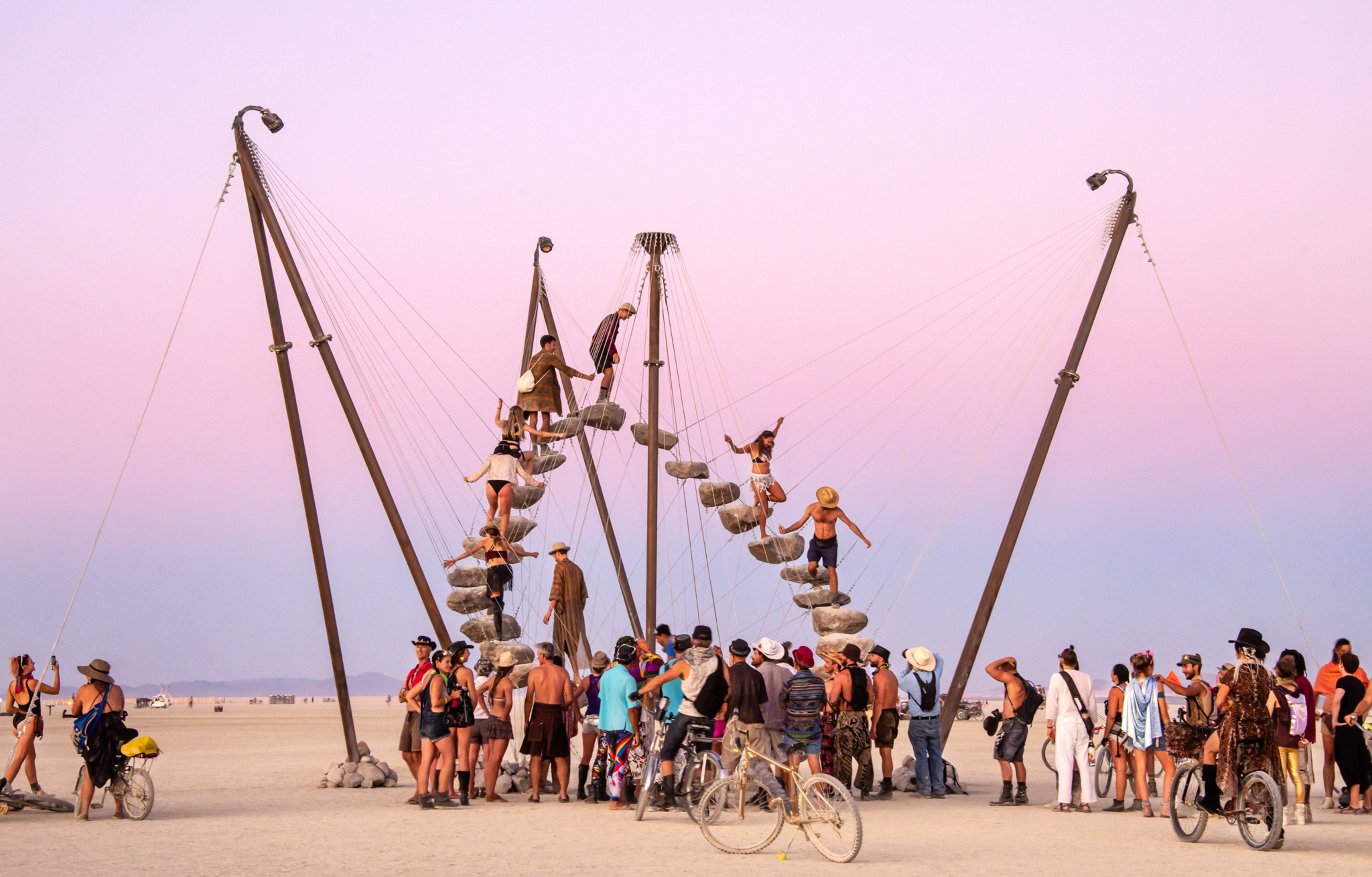 A group of people gather around and climb a circular art installation made of steps suspended in the air against a colorful sunset sky.