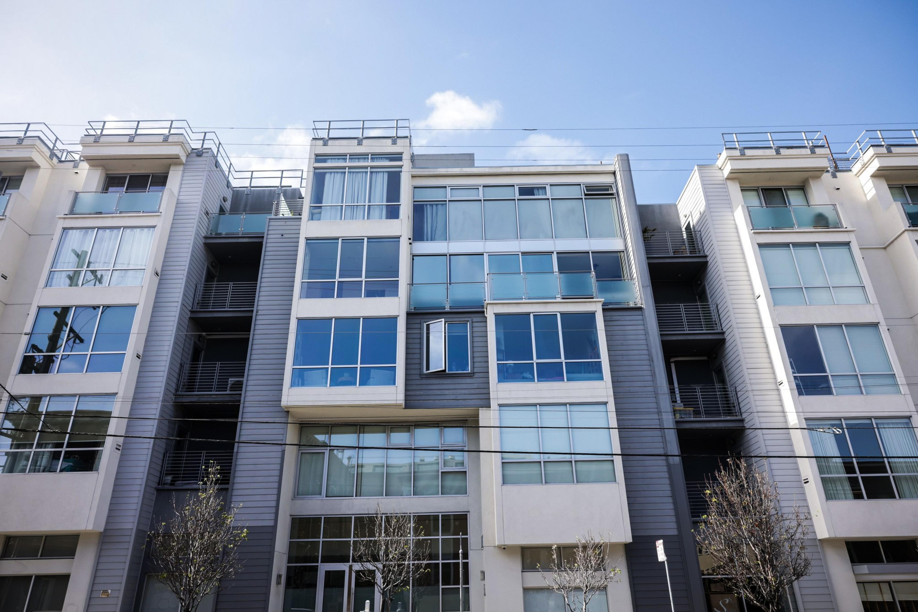 The image shows a modern apartment building with large glass windows, balconies, and a mix of grey and white exterior panels. Trees without leaves are in front.