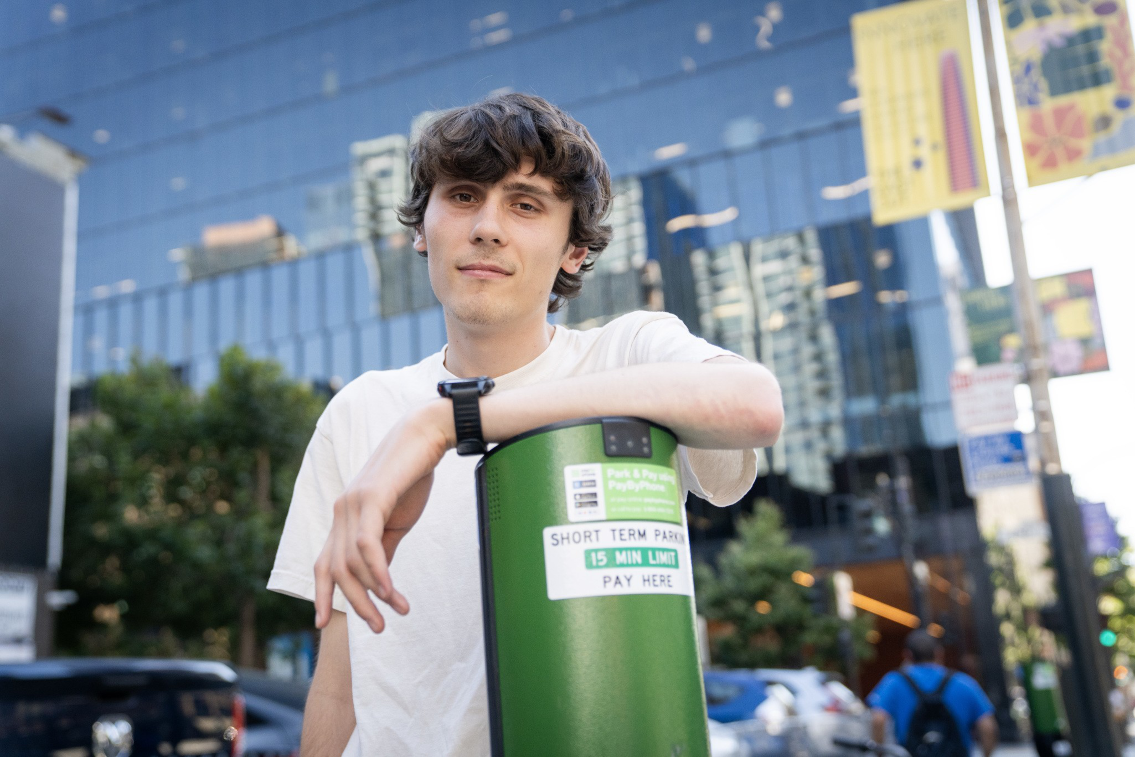 A young man in a white shirt leans on a green parking meter labeled “Short Term Park 15 Min Limit Pay Here” with urban buildings in the background.