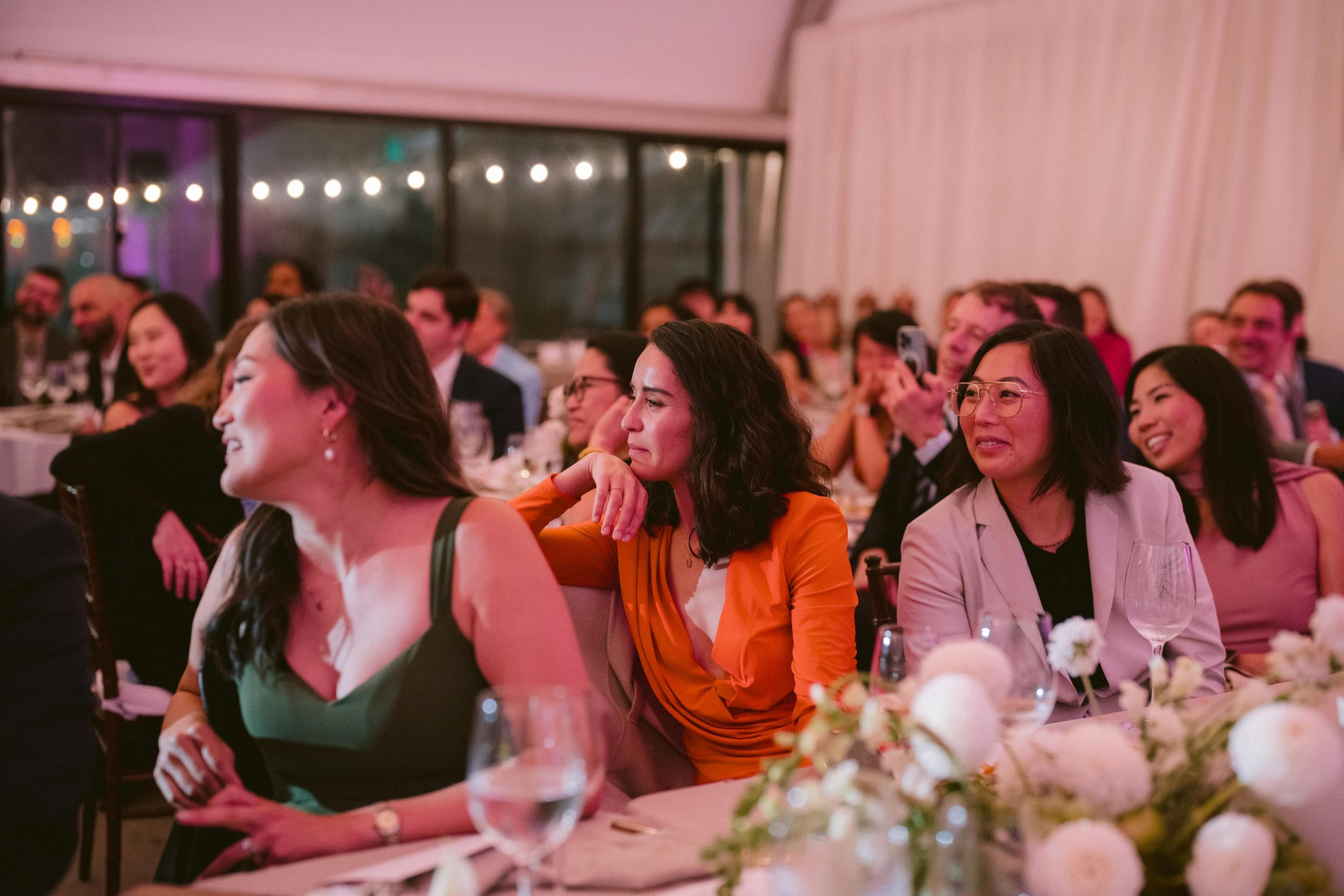 People dressed in formal attire sit at decorated tables, attentively watching something off-camera with smiles and focused expressions.