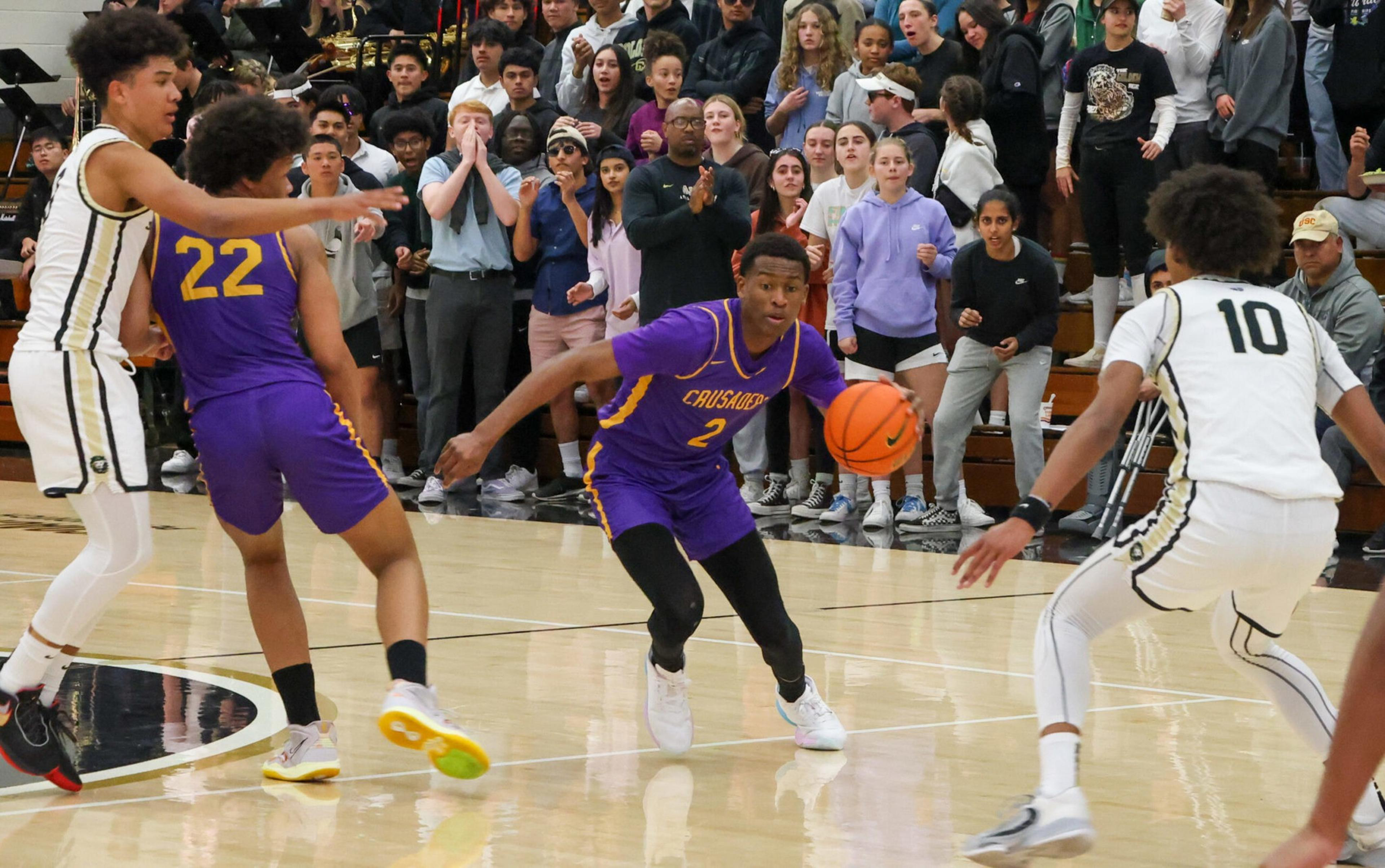 A basketball player in a purple jersey dribbles the ball while being closely guarded by players in white jerseys, with a crowd watching intently.