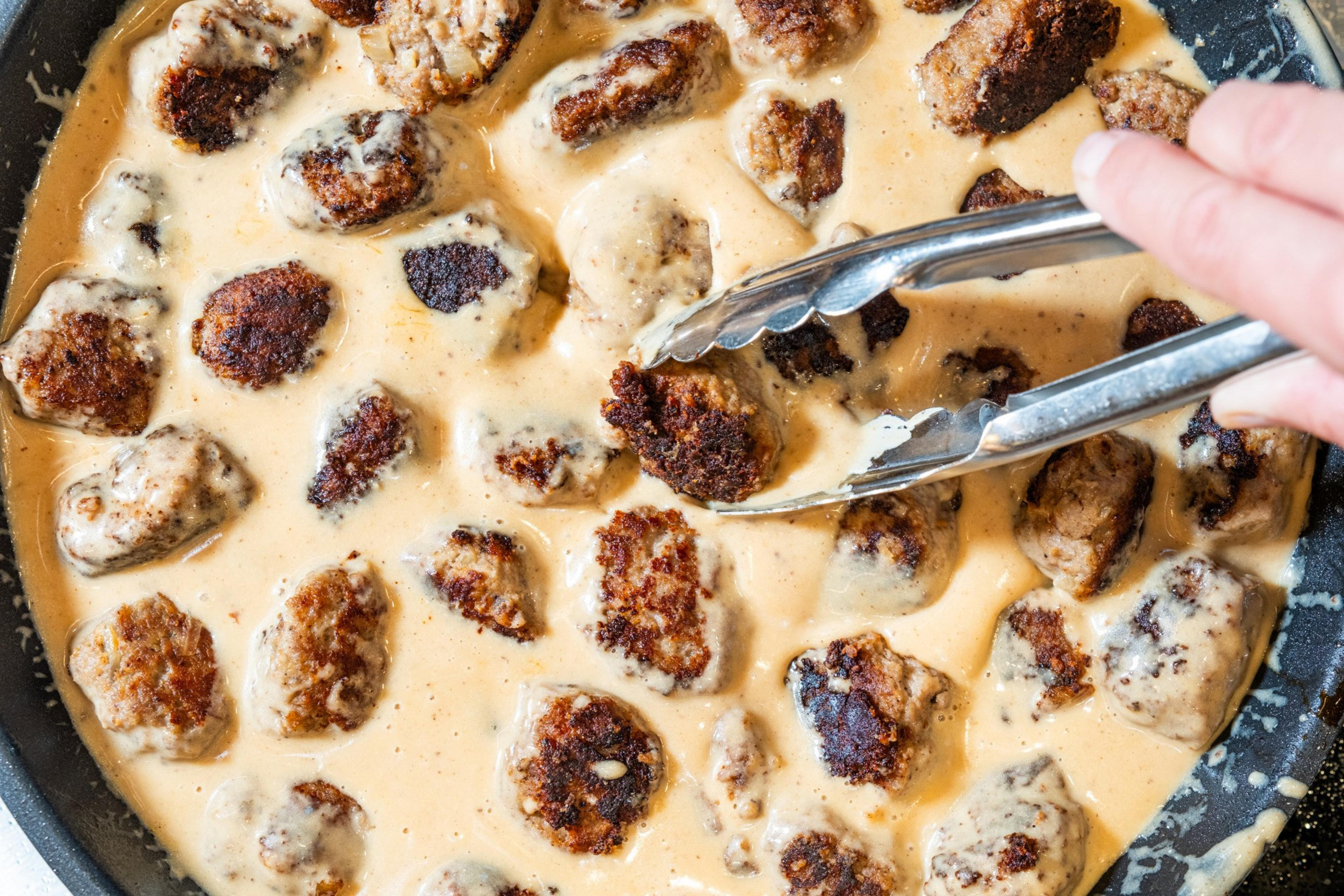 A pan of browned meatballs is submerged in a creamy sauce. A hand uses tongs to handle the meatballs, which appear to be coated in the sauce.