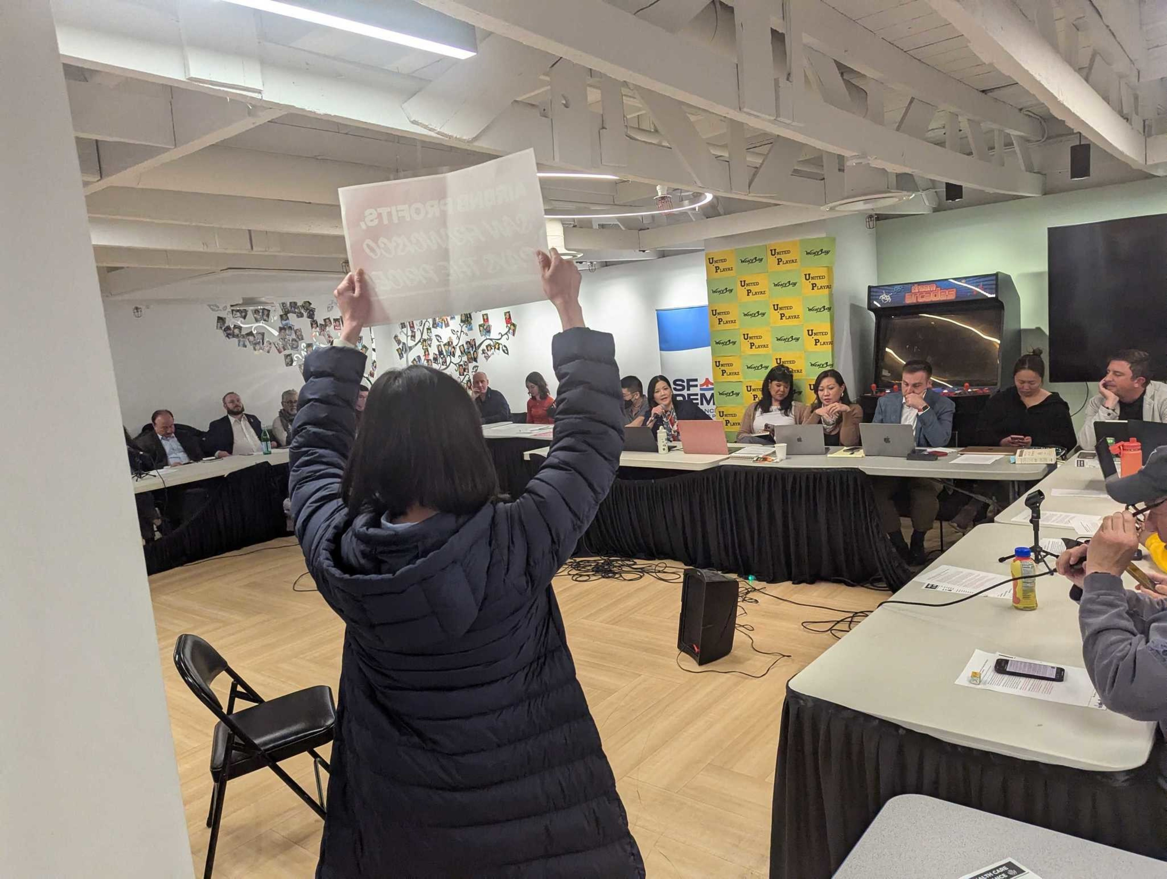 A woman in a black puffy jacket holds a protest sign aloft, reading &quot;Airbnb profits, San Francisco pays the price!&quot; Shhe is seen from behind, with Democratic Party board members sitting along a series of white tables in the background.