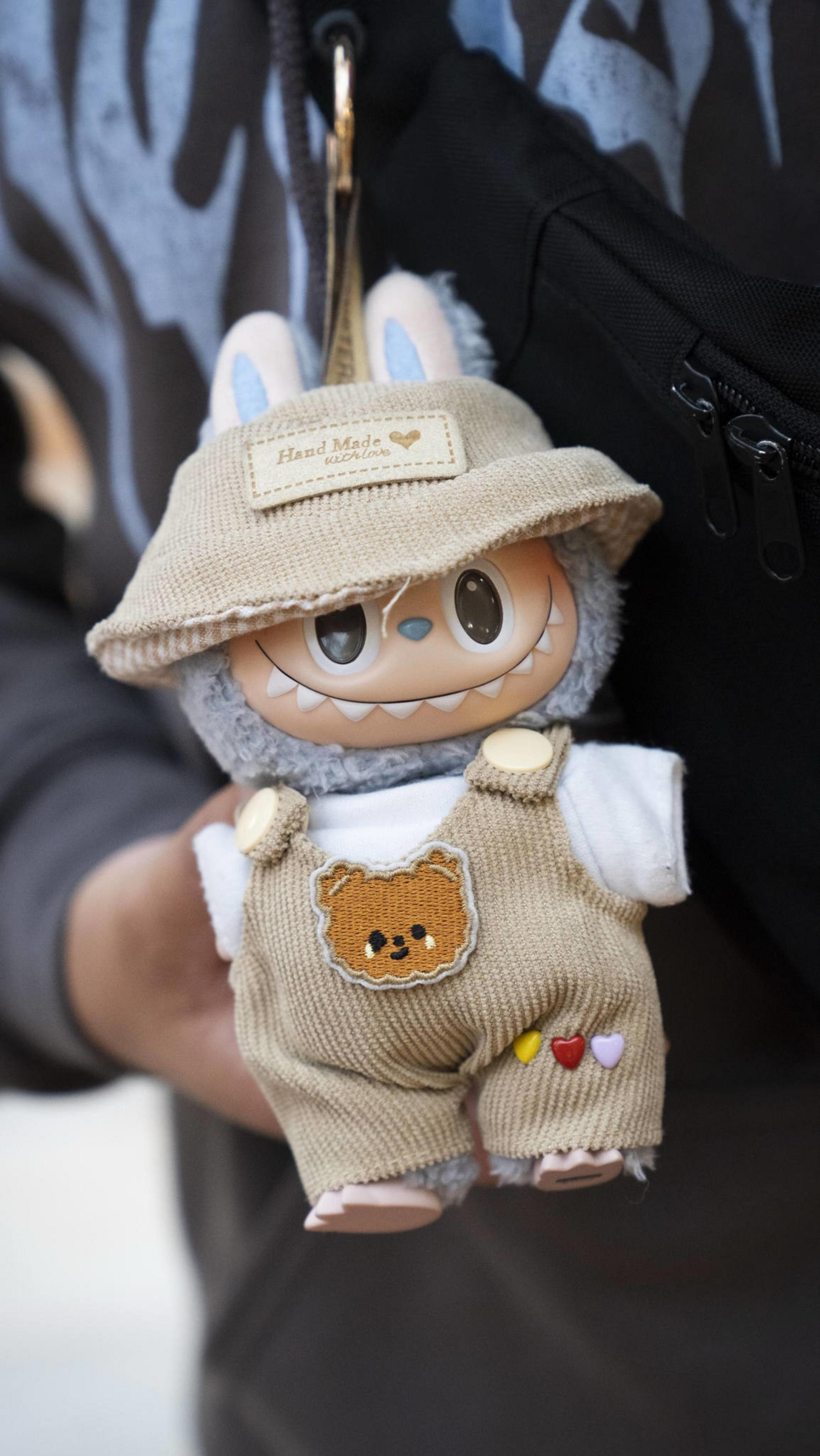 A plush toy with a cute, grinning face, wearing a beige bucket hat and overalls featuring a bear patch and small heart buttons, held in someone's hand.