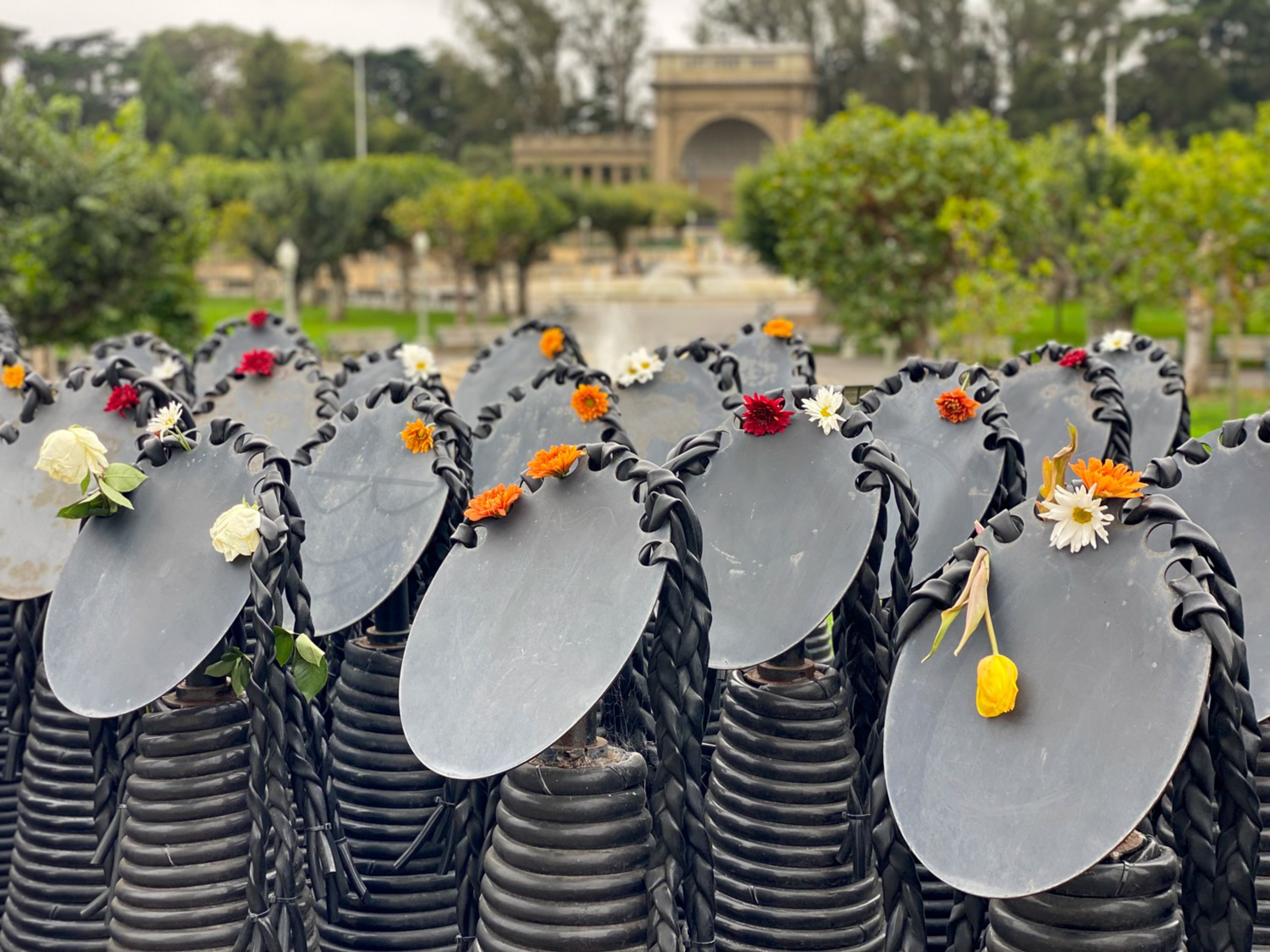 Rows of dark, helmet-shaped sculptures with round shields decorated with colorful flowers stand in an outdoor garden setting.