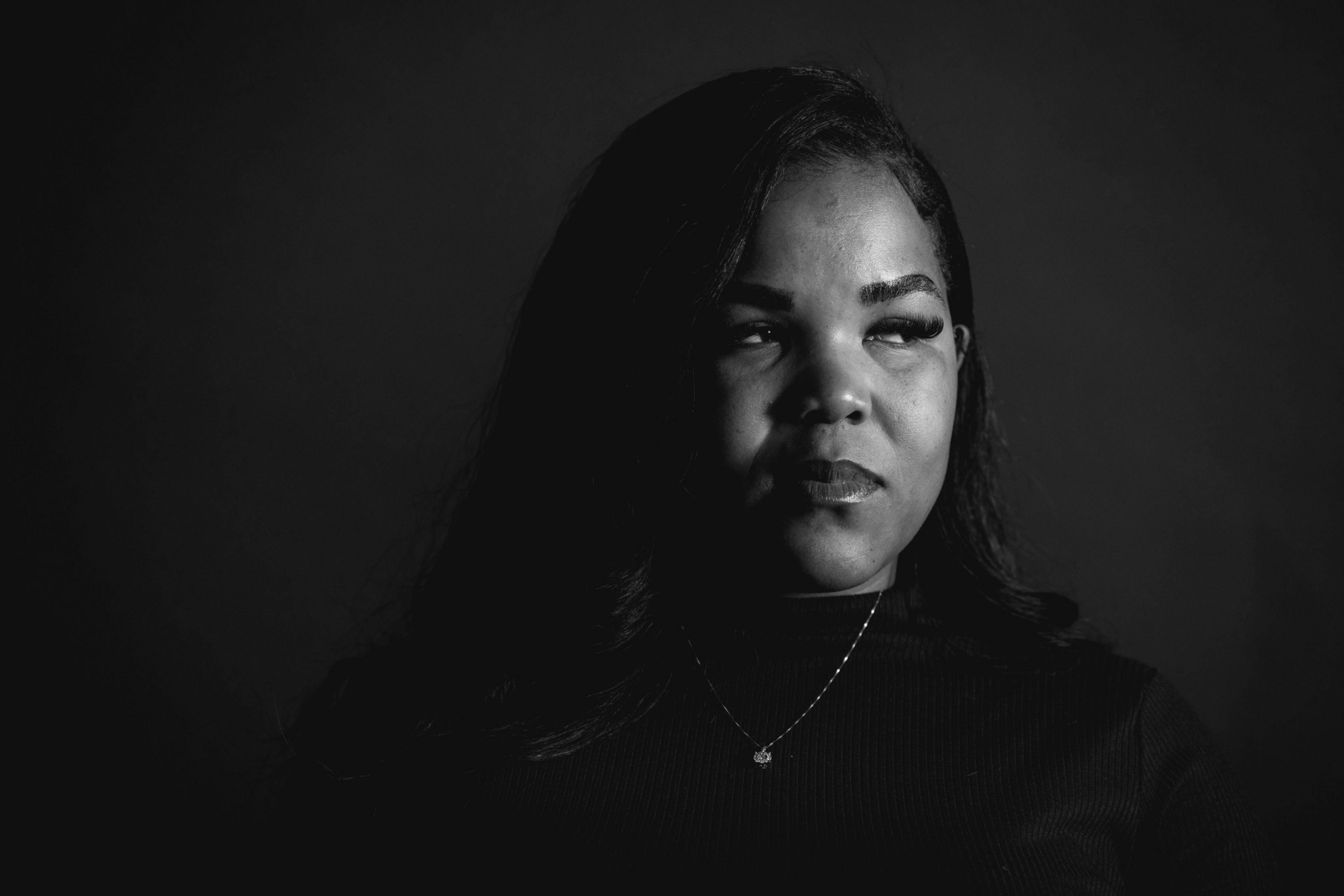 A black-and-white portrait of a person with long dark hair, looking slightly to the side. They are wearing a necklace and a dark top, set against a dark background.