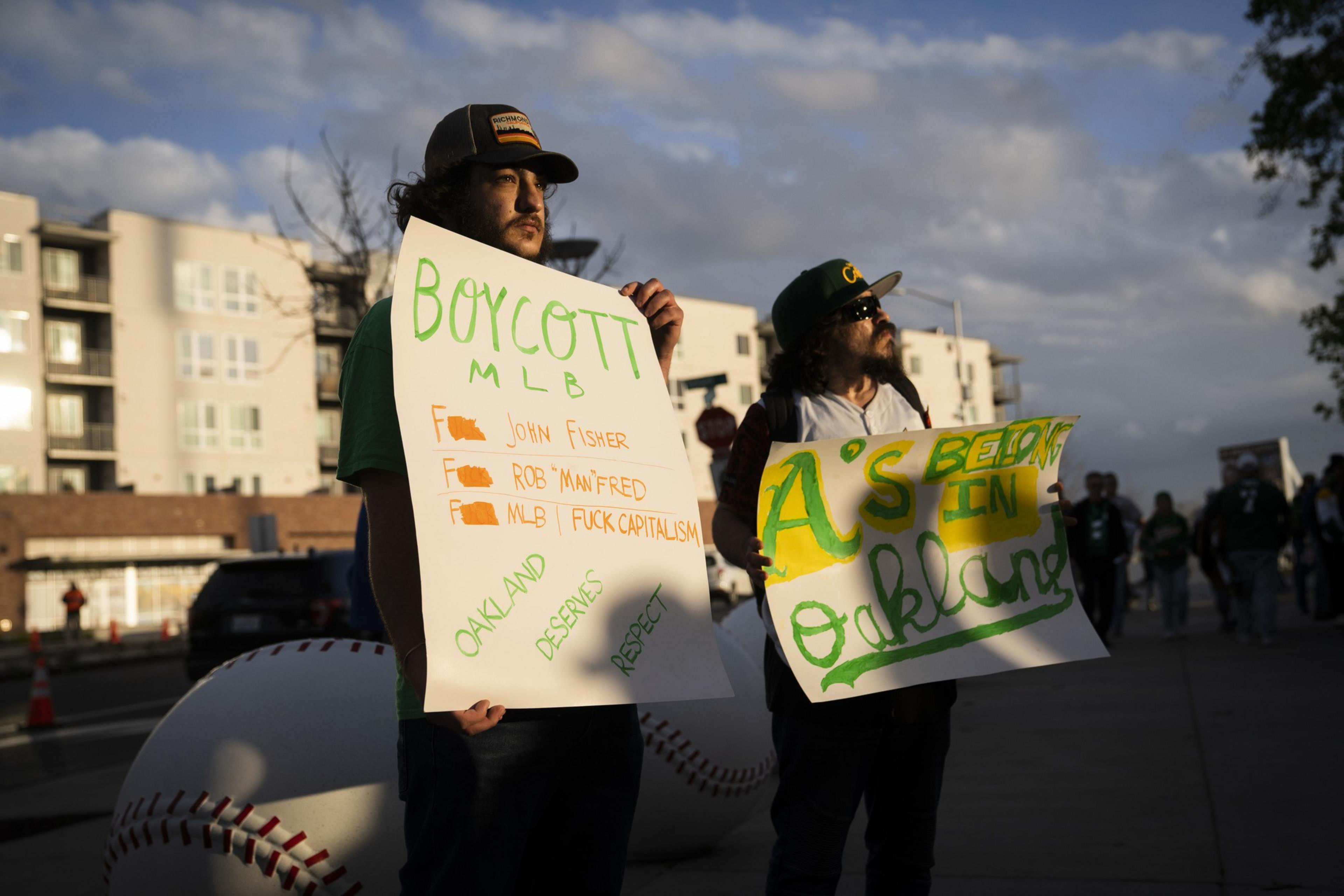 Two people hold protest signs. One reads &quot;Boycott MLB,&quot; listing grievances, and the other says &quot;A's Belong in Oakland.&quot; They're standing on a street.