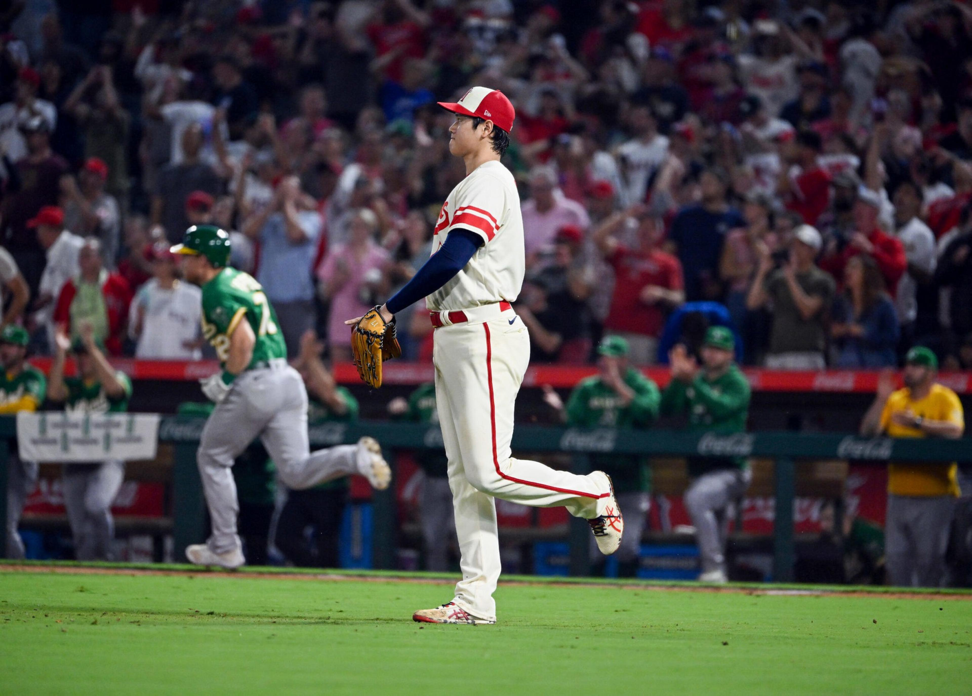 A baseball player in white stands on the field with one leg raised, while a player in green runs past and the crowd cheers in the background.