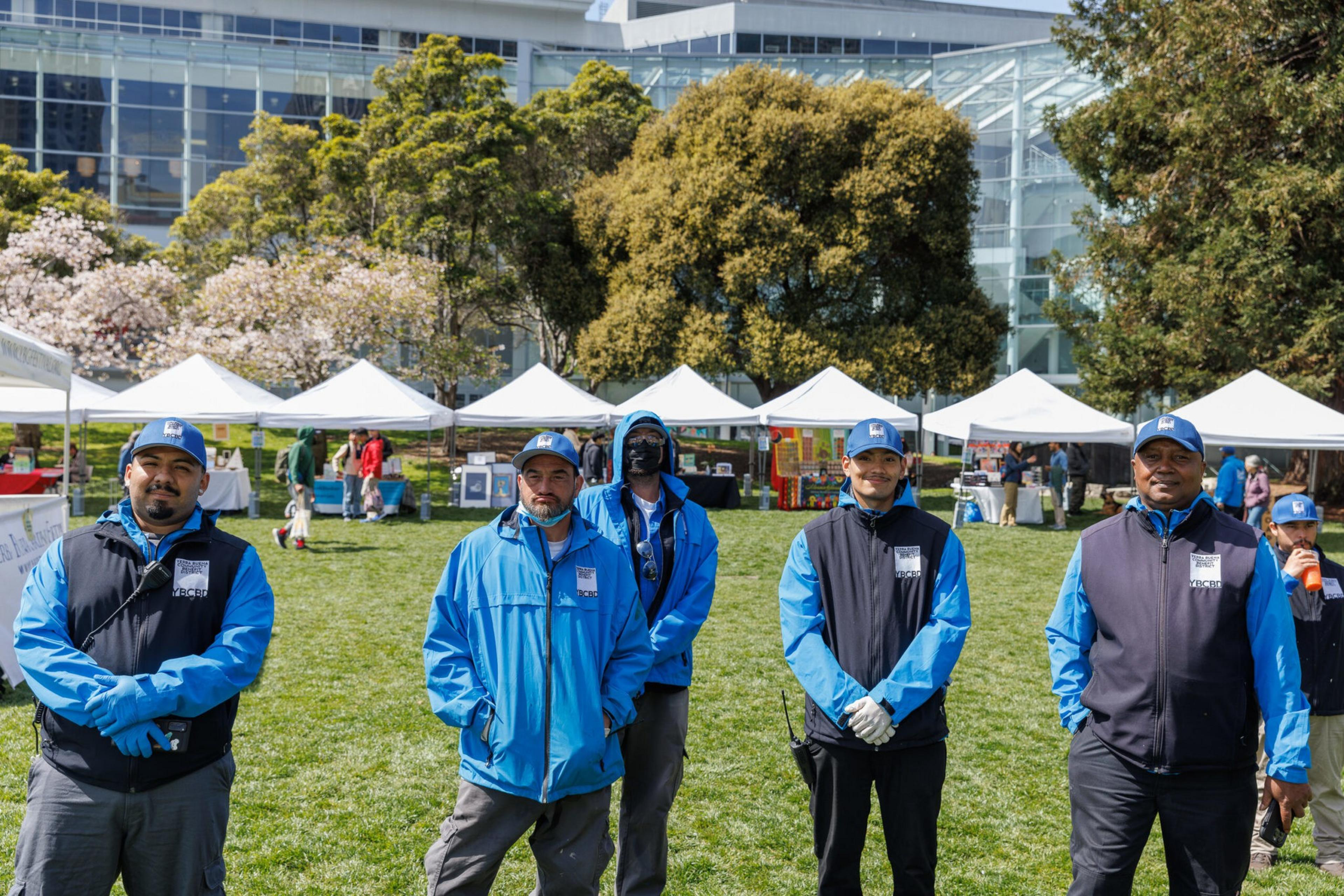 Six event staff in blue jackets stand on a green lawn with event tents and a modern building behind them.