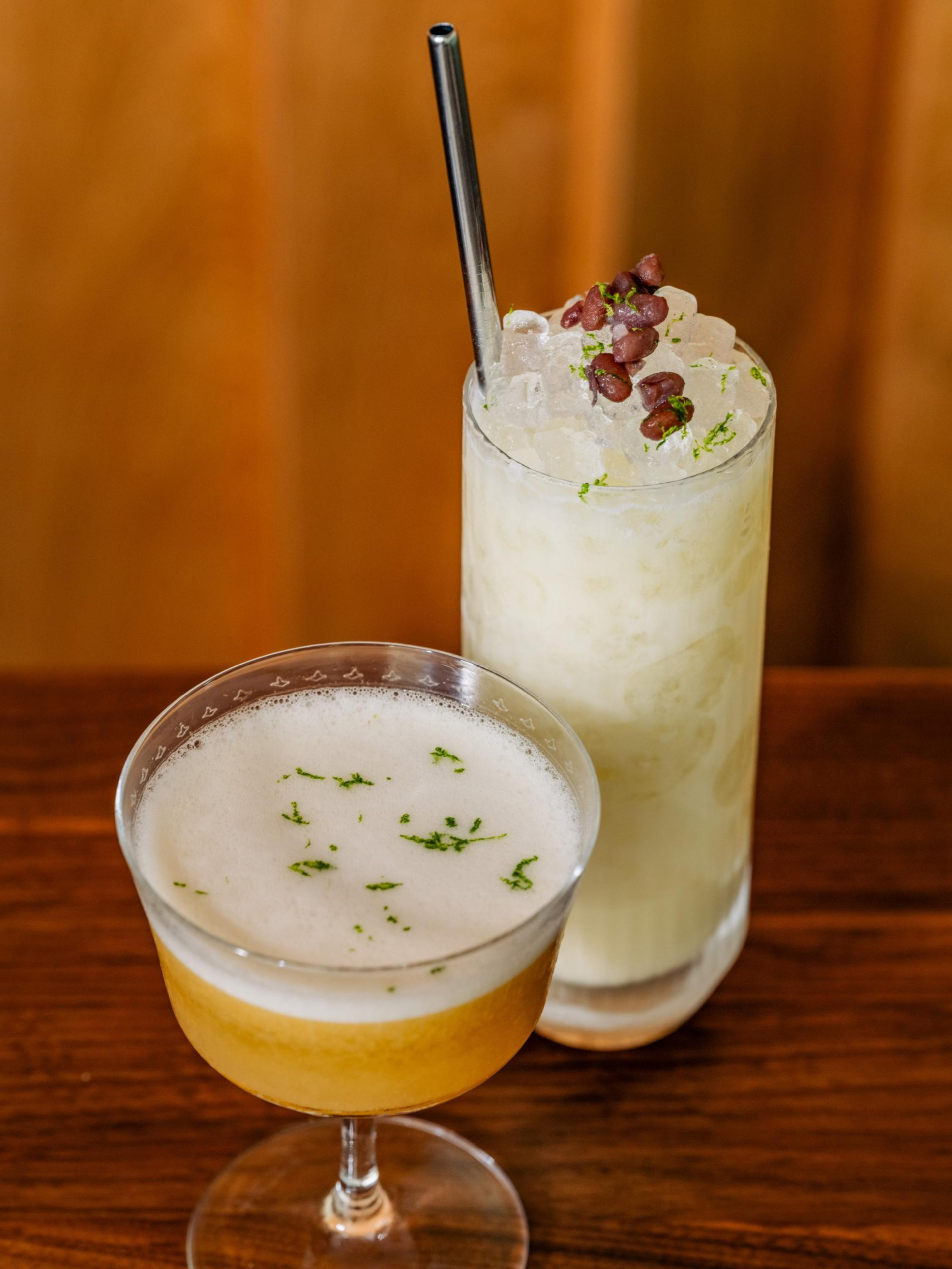 A foamy yellow cocktail garnished with lime zest sits next to a tall iced white drink topped with red beans and a metal straw on a wooden table.