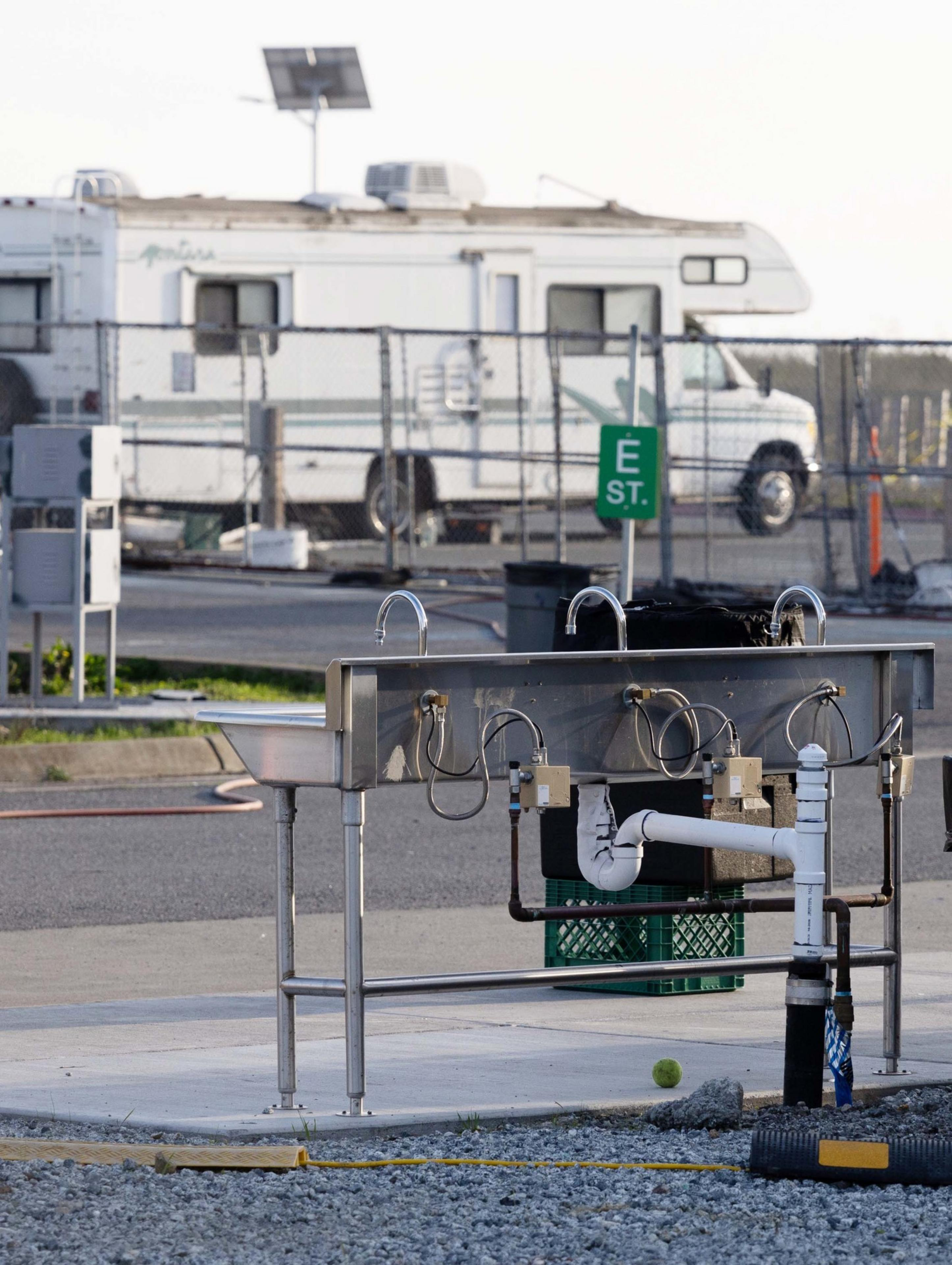 The image shows a silver outdoor washing station with multiple faucets, set on concrete near a gravel area. An RV and fenced area are visible in the background.