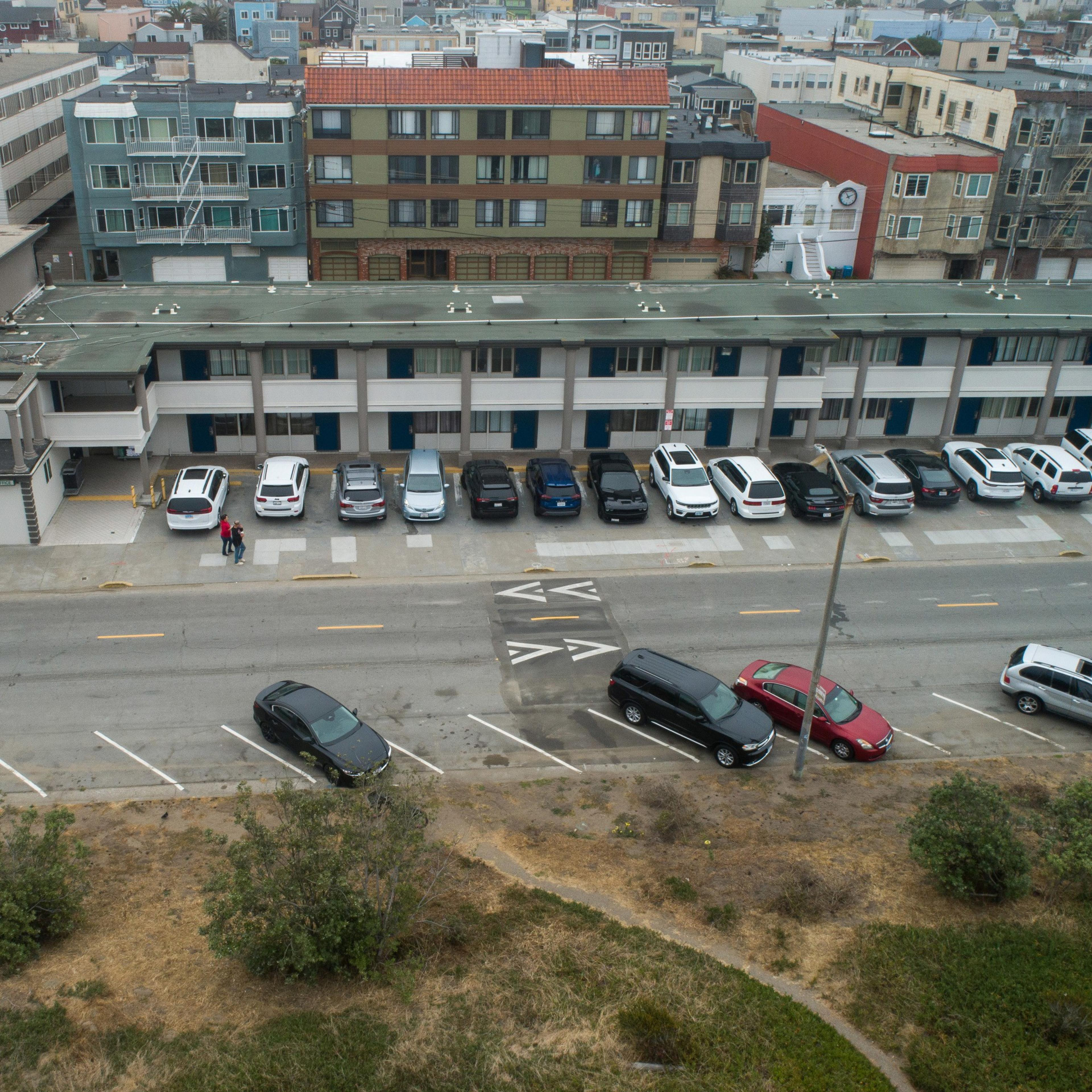 The image depicts a street scene with a parking lot where multiple cars are parked in front of a multi-story apartment building. Two people stand on the sidewalk.