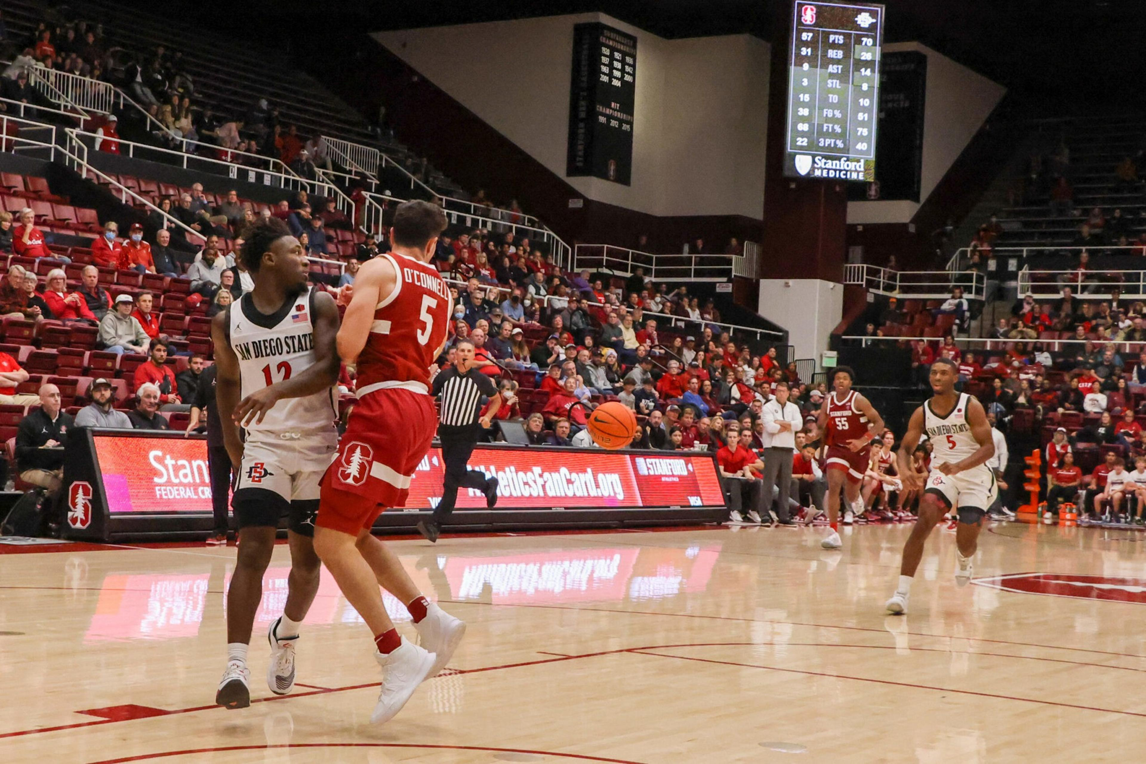 Two basketball players, one in white and one in red, compete near the sideline while another white team player runs toward the ball on a packed court.