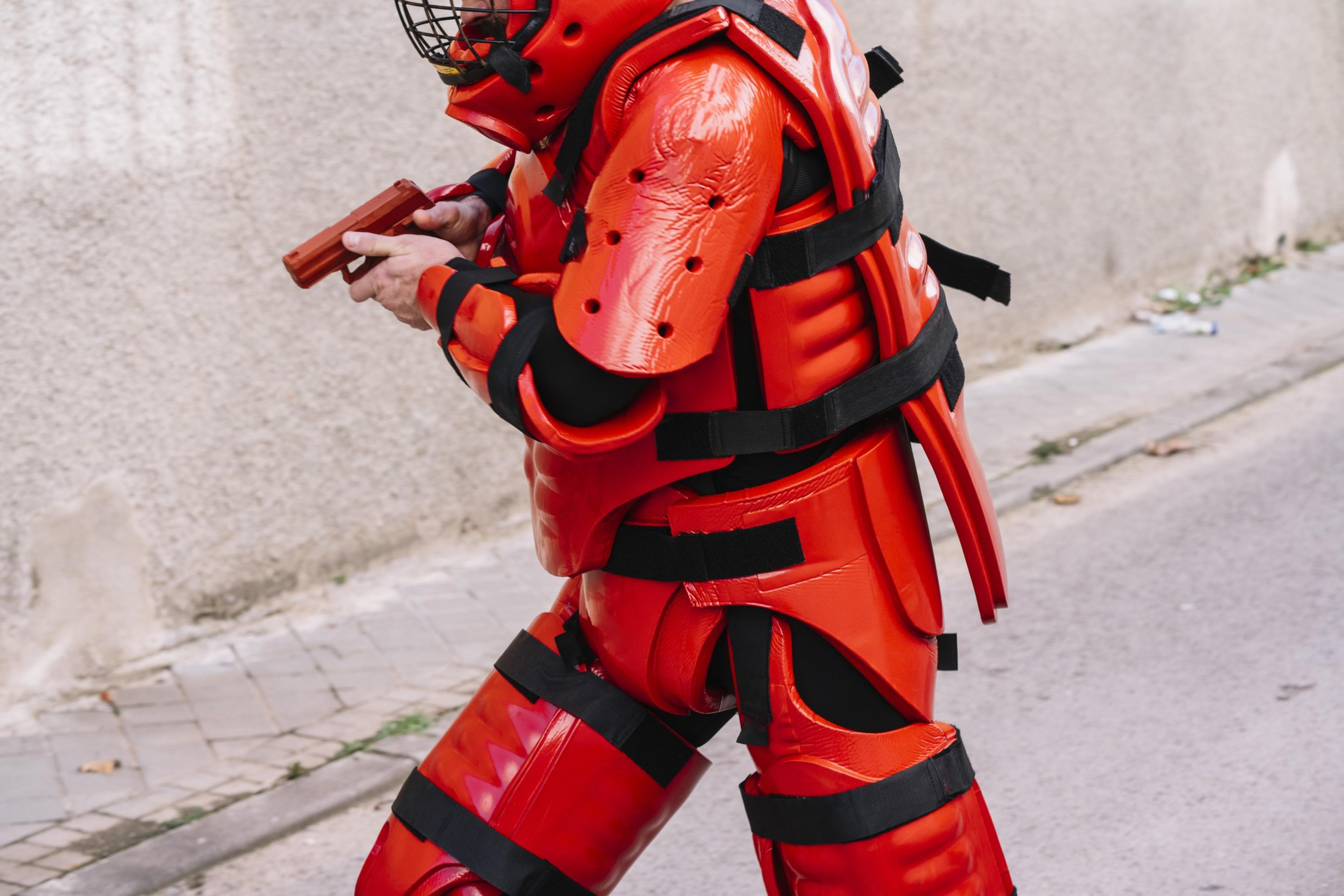 A person dressed in red protective gear and helmet, holding a red plastic gun, is crouching on a street next to a concrete wall.