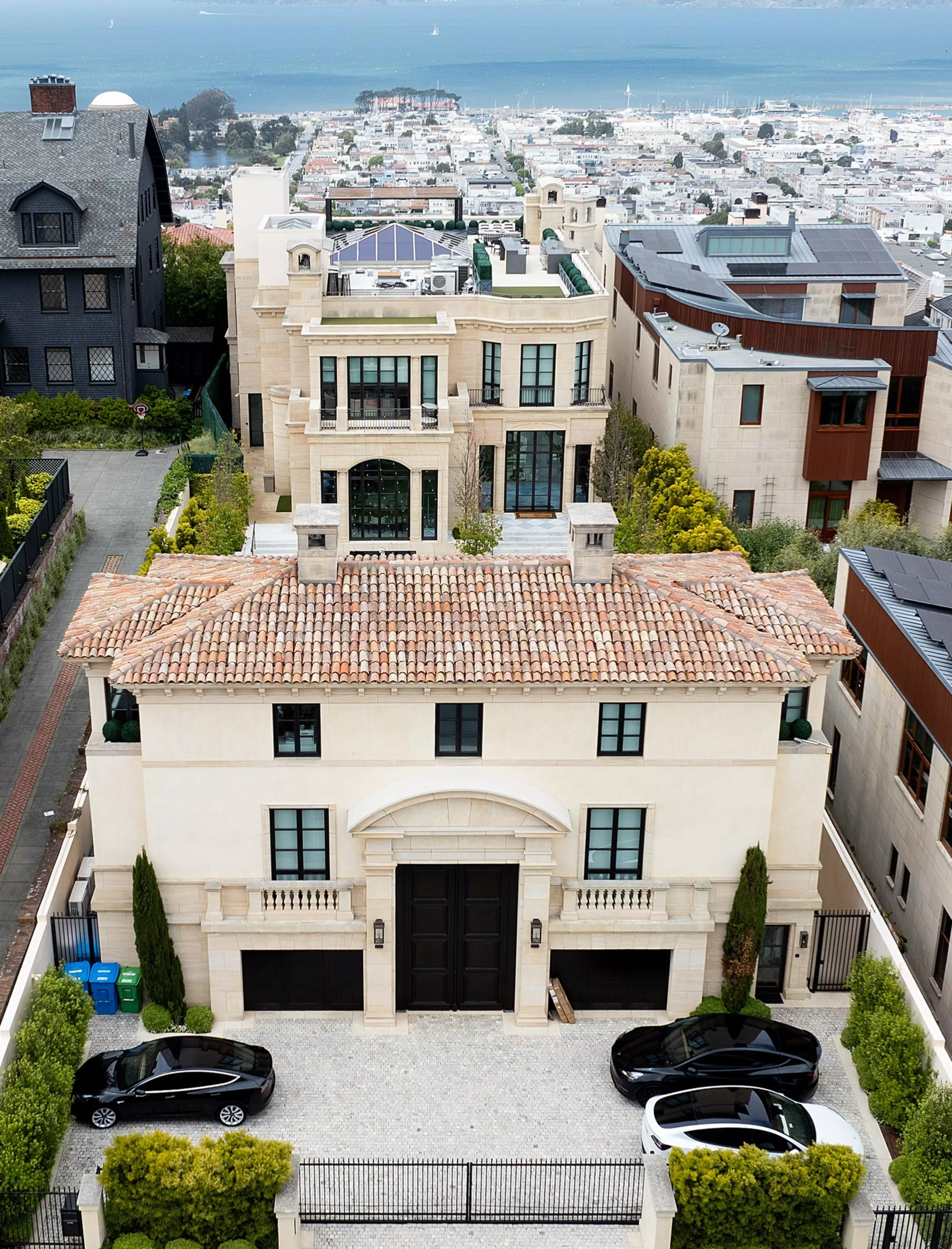 A large, luxurious house with a tile roof and gated driveway containing two black cars sits in a hilly neighborhood. Behind it is a cityscape and a body of water.